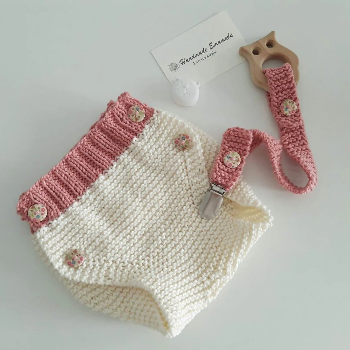 Culotte a maglia bicolore panna e rosa con bottoni a fiori, accanto a un portaciuccio a maglia rosa con anello in legno a gufo.