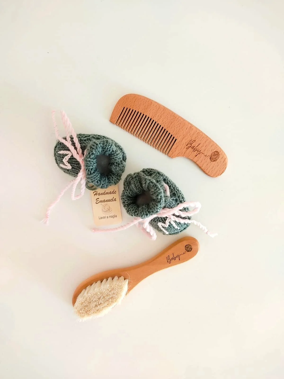 Pettinino e spazzolina in legno con incisione Baby accanto a un paio di scarpine in maglia verdi con cartellino