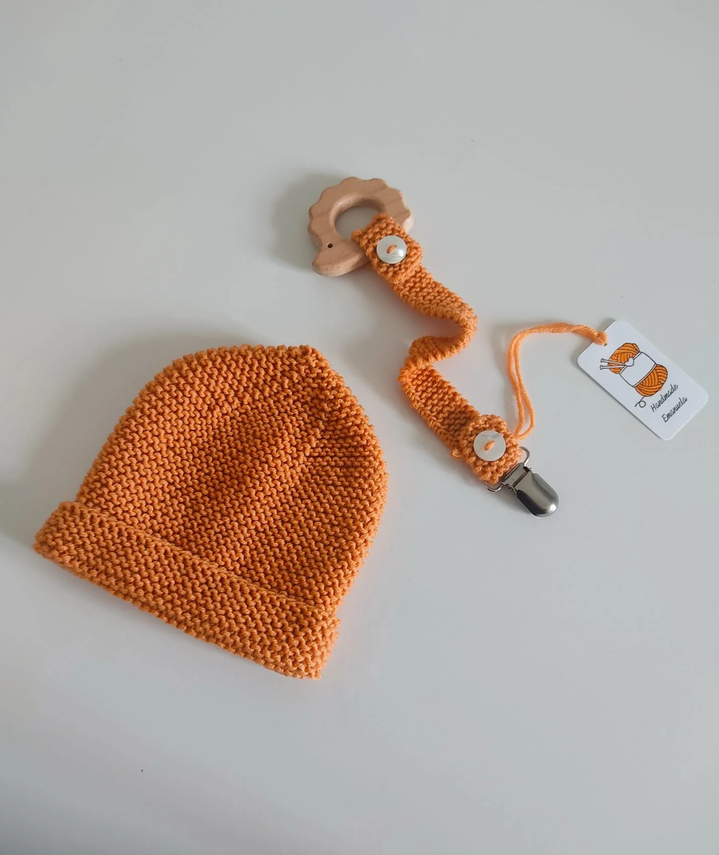 Portaciuccio in maglia arancione con giochino in legno a riccio, accanto a un cappellino arancione, con cartellino