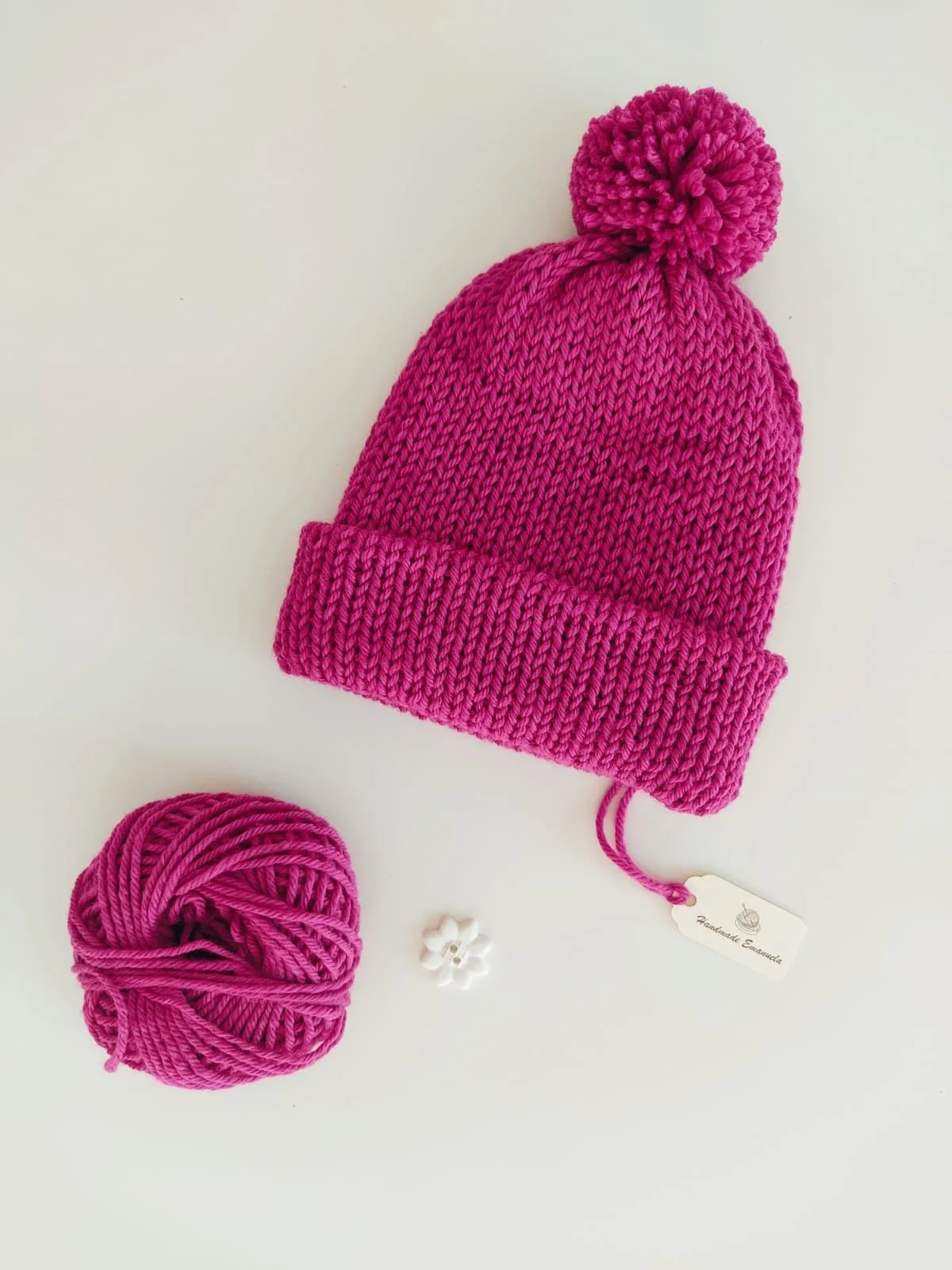 Cappello in maglia fucsia con risvolto e pompon dello stesso colore, accanto a un gomitolo, con cartellino