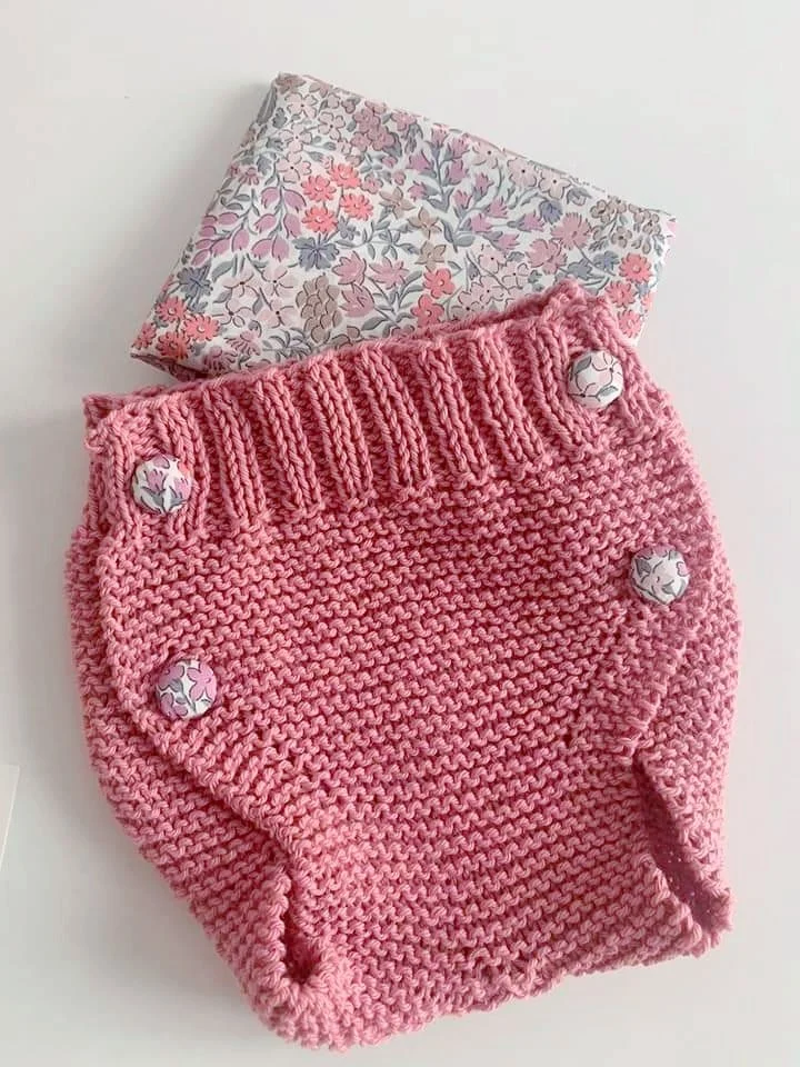 Culotte a maglia rosa con bottoni rivestiti in stoffa a fiori, appoggiata su un tessuto floreale rosa e grigio.
