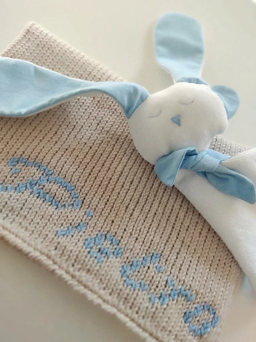 Doudou coniglietto in spugna bianca con orecchie azzurre, su una copertina in maglia beige con una scritta azzurra