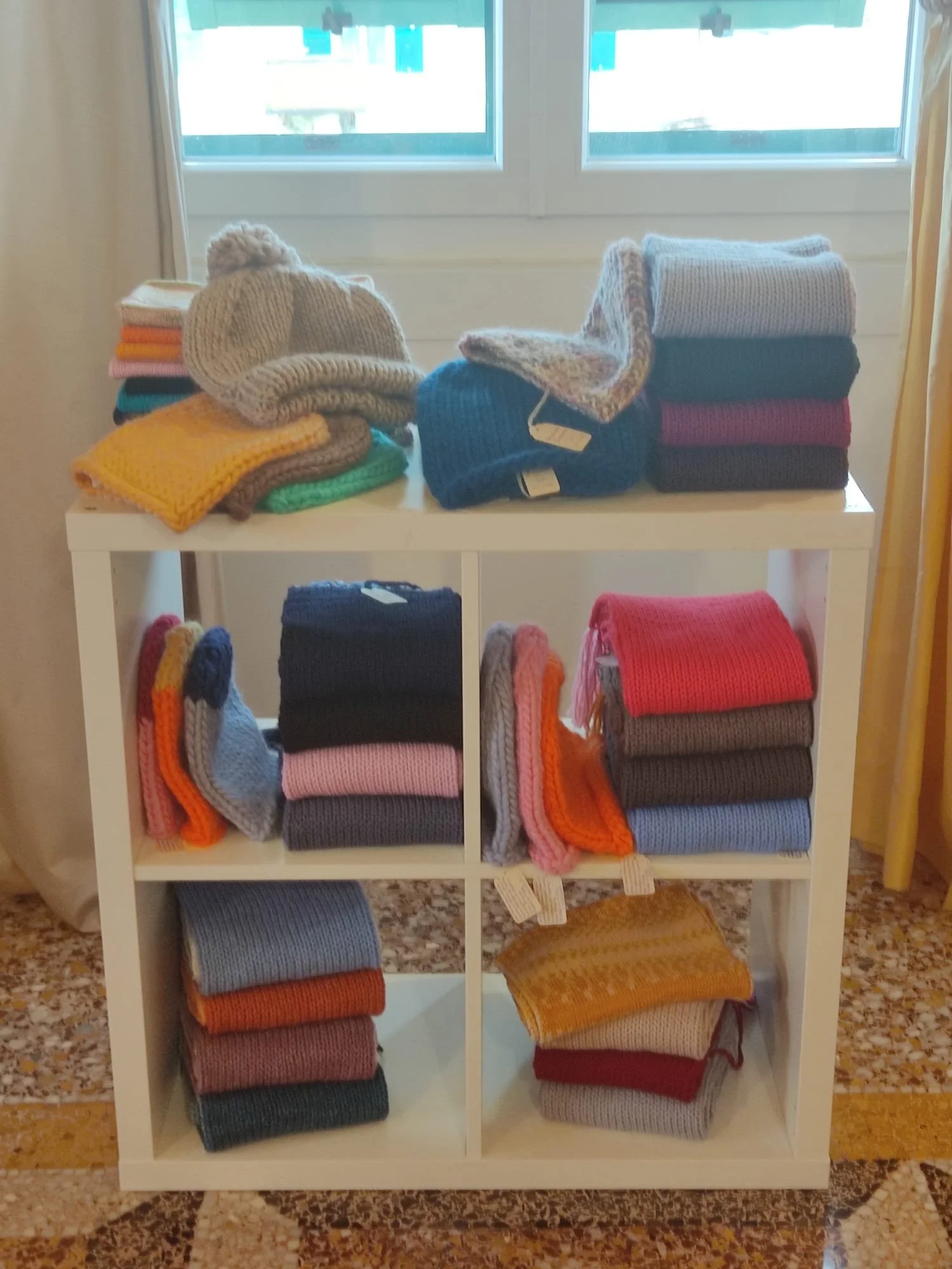 Sciarpe e cappelli in maglia di lana piegati su uno scaffale bianco, in tanti colori vivaci, con cartellini appesi