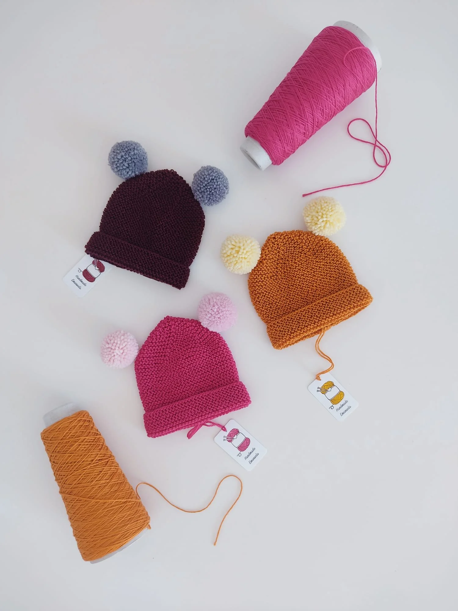 Quattro cappelli in maglia con risvolto e due pompon, bordeaux, fucsia e arancione, accanto a due rocche di filo