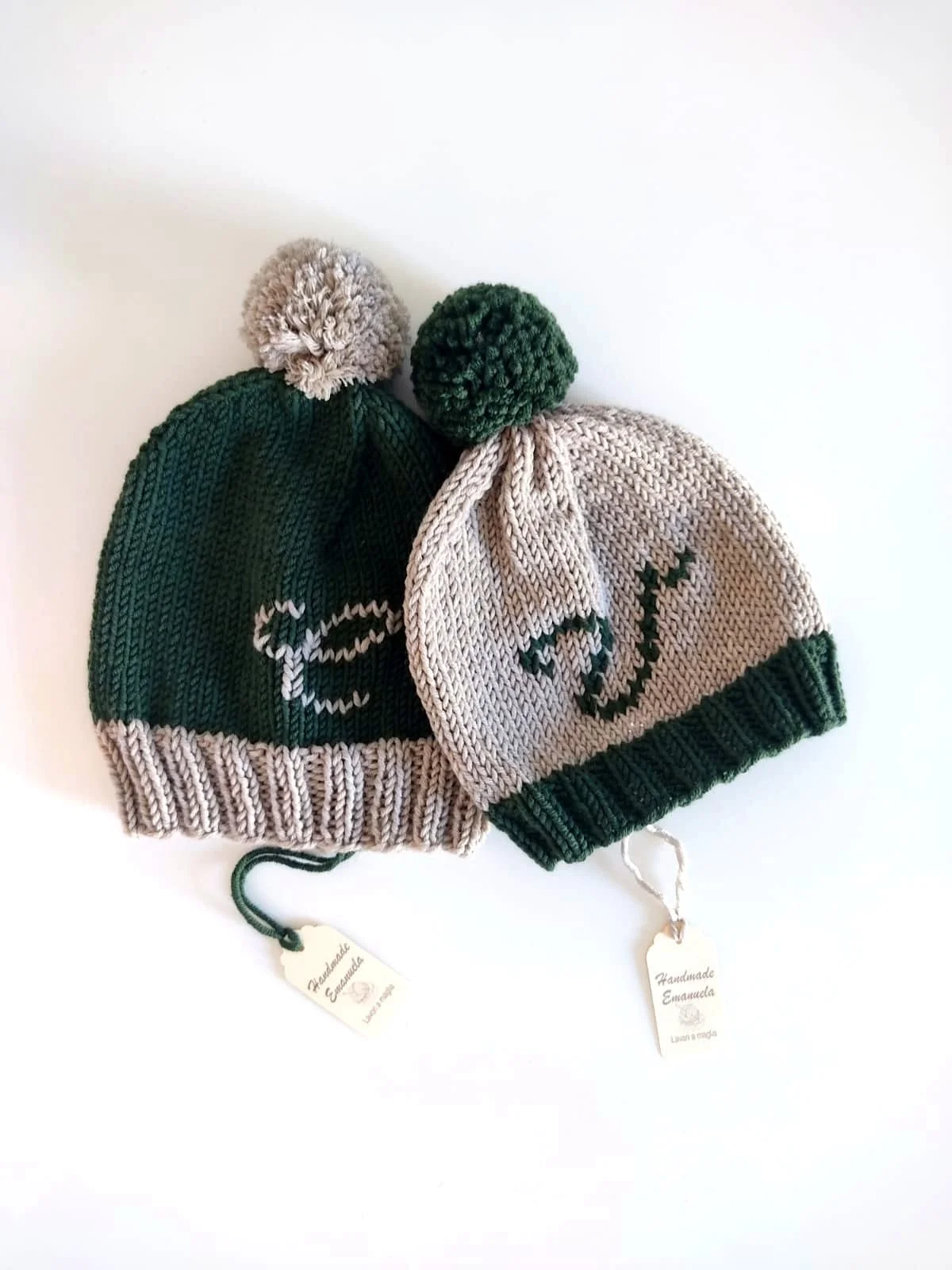 Due cappelli in maglia verde scuro e beige con pompon, sovrapposti, con iniziale ricamata e cartellino