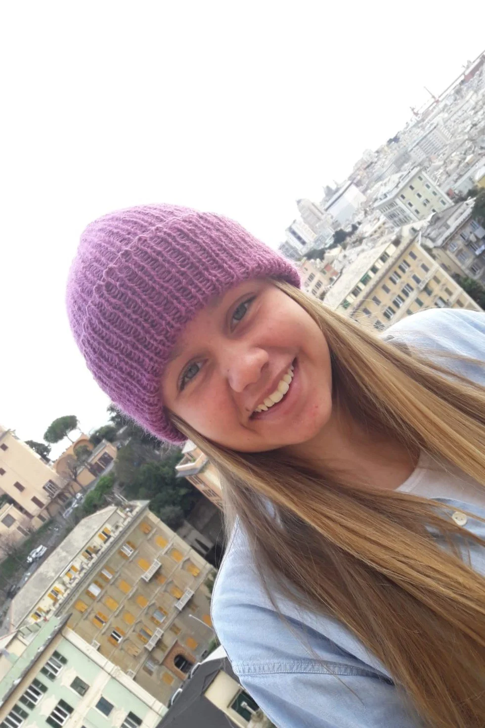 Ragazza che sorride con un cappello in maglia lilla a coste, ripresa in diagonale con case sullo sfondo