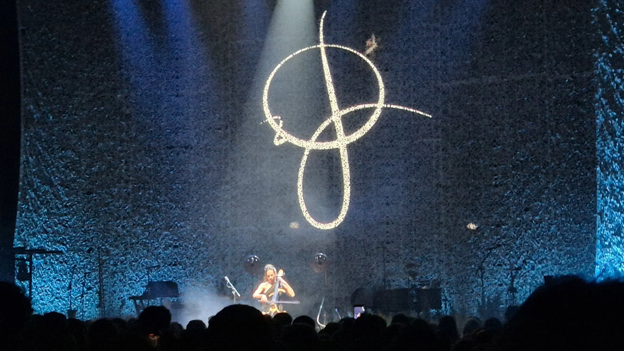 A stage with a large peace sign symbol outlined with lights hanging above a musician playing a cello. The stage has dark textured walls and blue lighting.