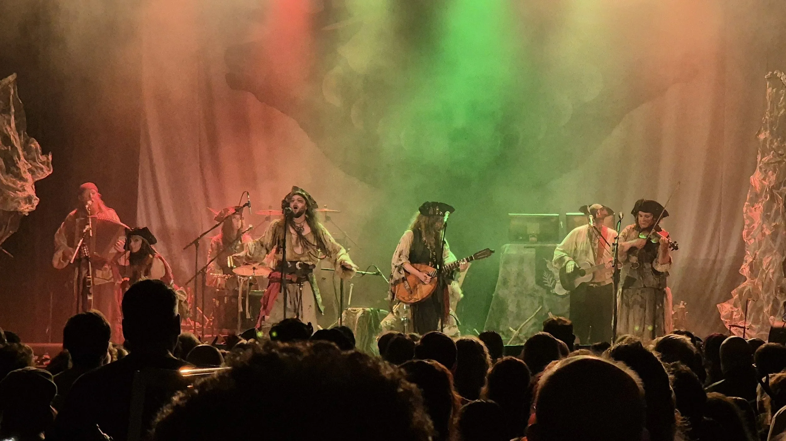 A live musical performance on stage with musicians dressed in pirate costumes, playing instruments under green and orange stage lights, with a seated audience watching.