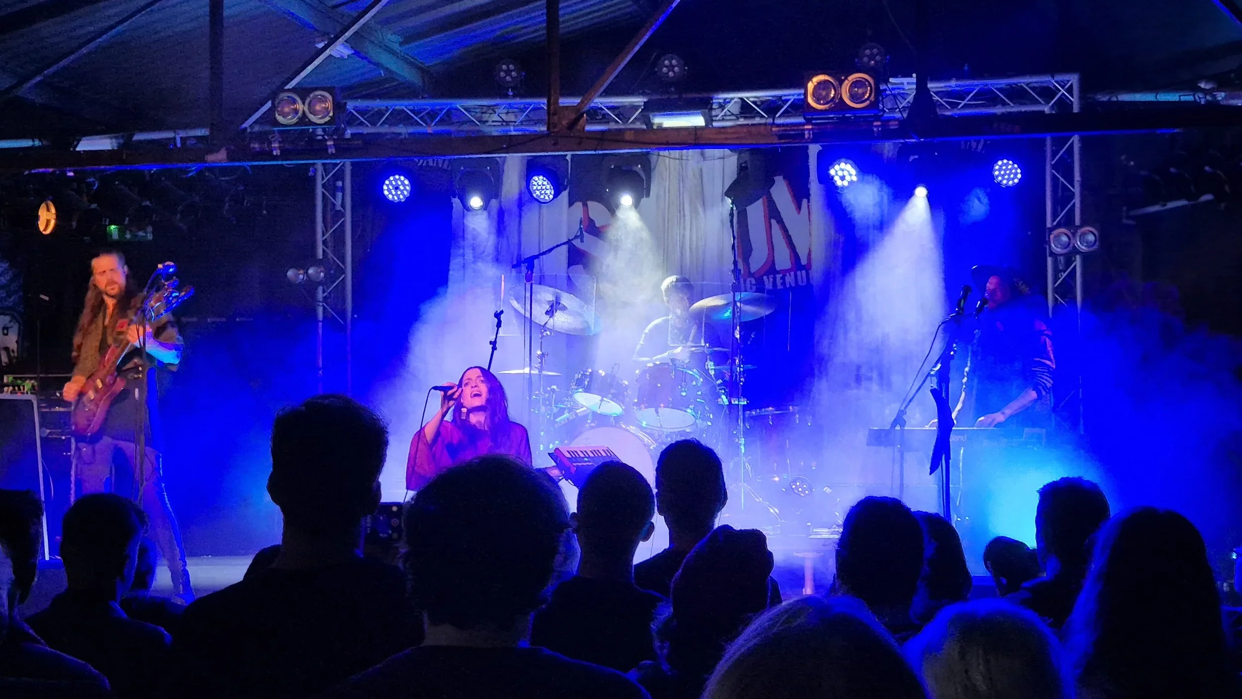 A live music performance with a band playing on stage. The singer is in the front, holding a microphone. The drummer is in the center, and there are two other musicians playing guitar and keyboard. The stage is lit with blue and purple lights, and th