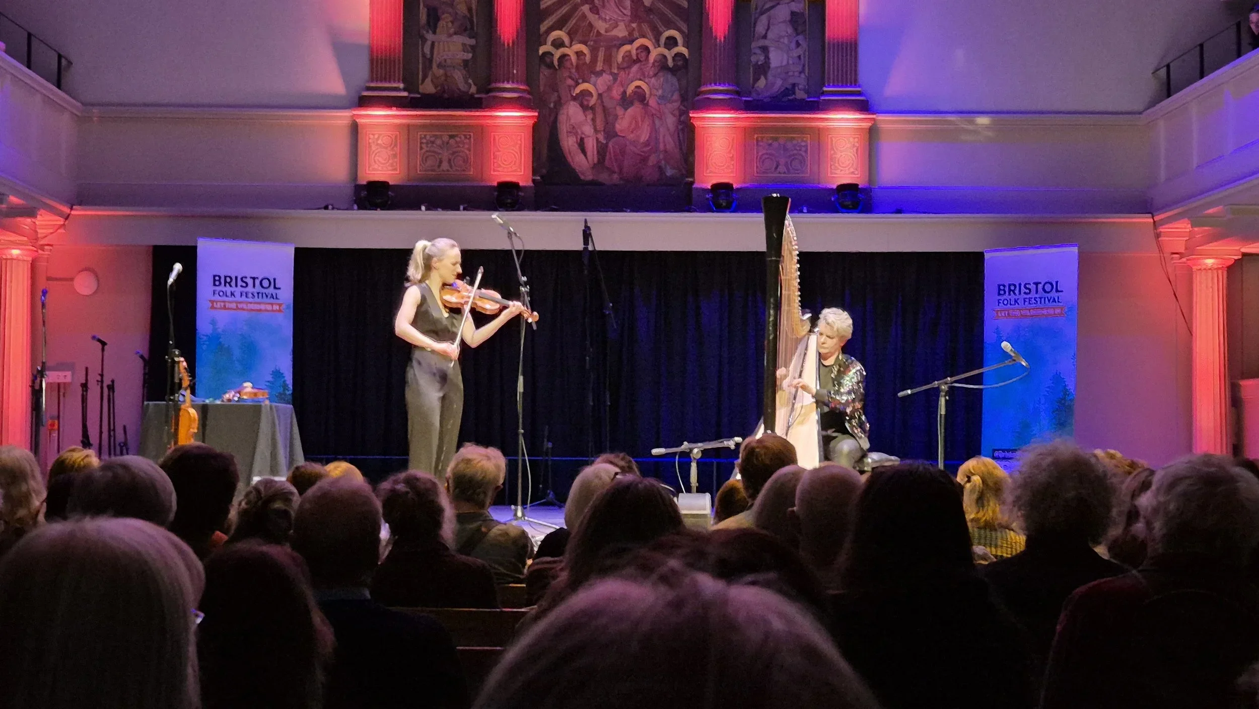 Two female musicians performing on stage at the Bristol Folk Festival, one playing a violin and the other on a harp, with an audience watching.