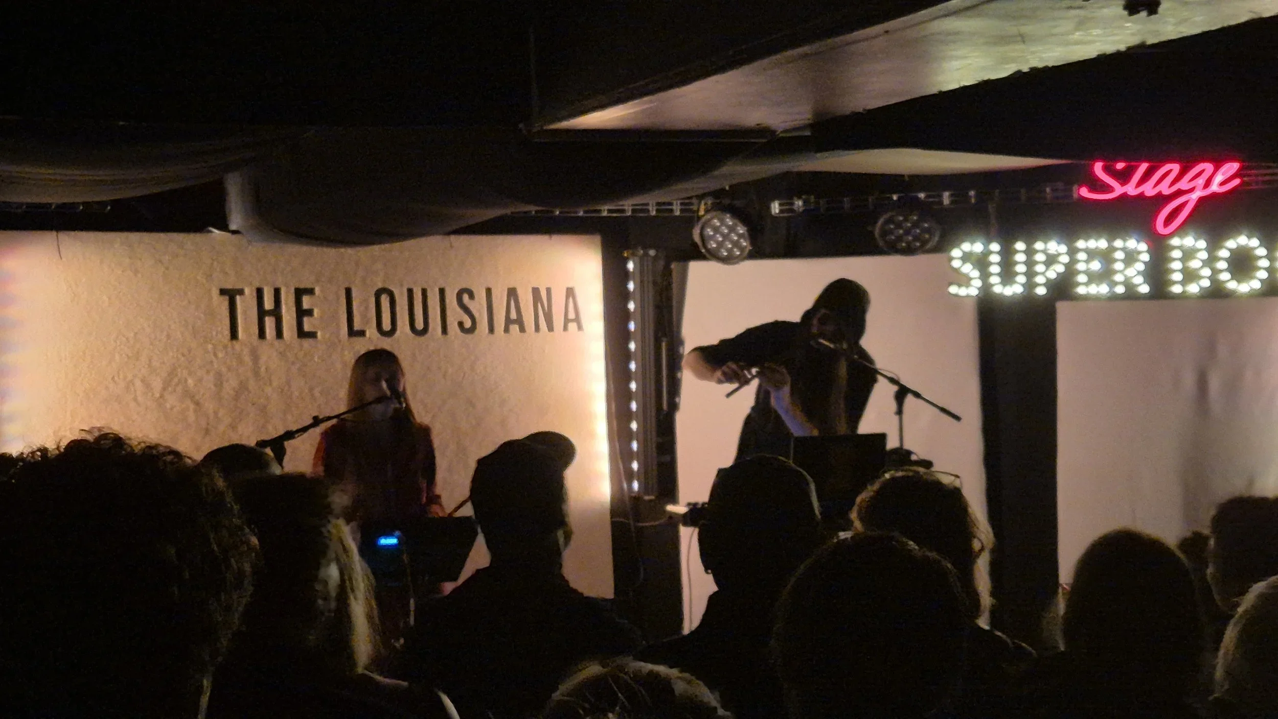A live performance at The Louisiana with musicians on stage, one playing guitar and another singing into a microphone, in a venue with dim lighting and audience members visible in the foreground. A neon sign reads 'Stage' and a digital sign displays 