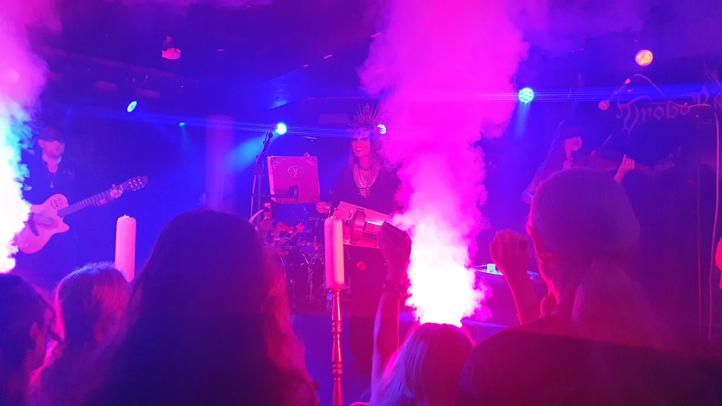 A live music band performing on stage with colorful purple and blue lighting, a smoke or fog machine, and an audience watching.