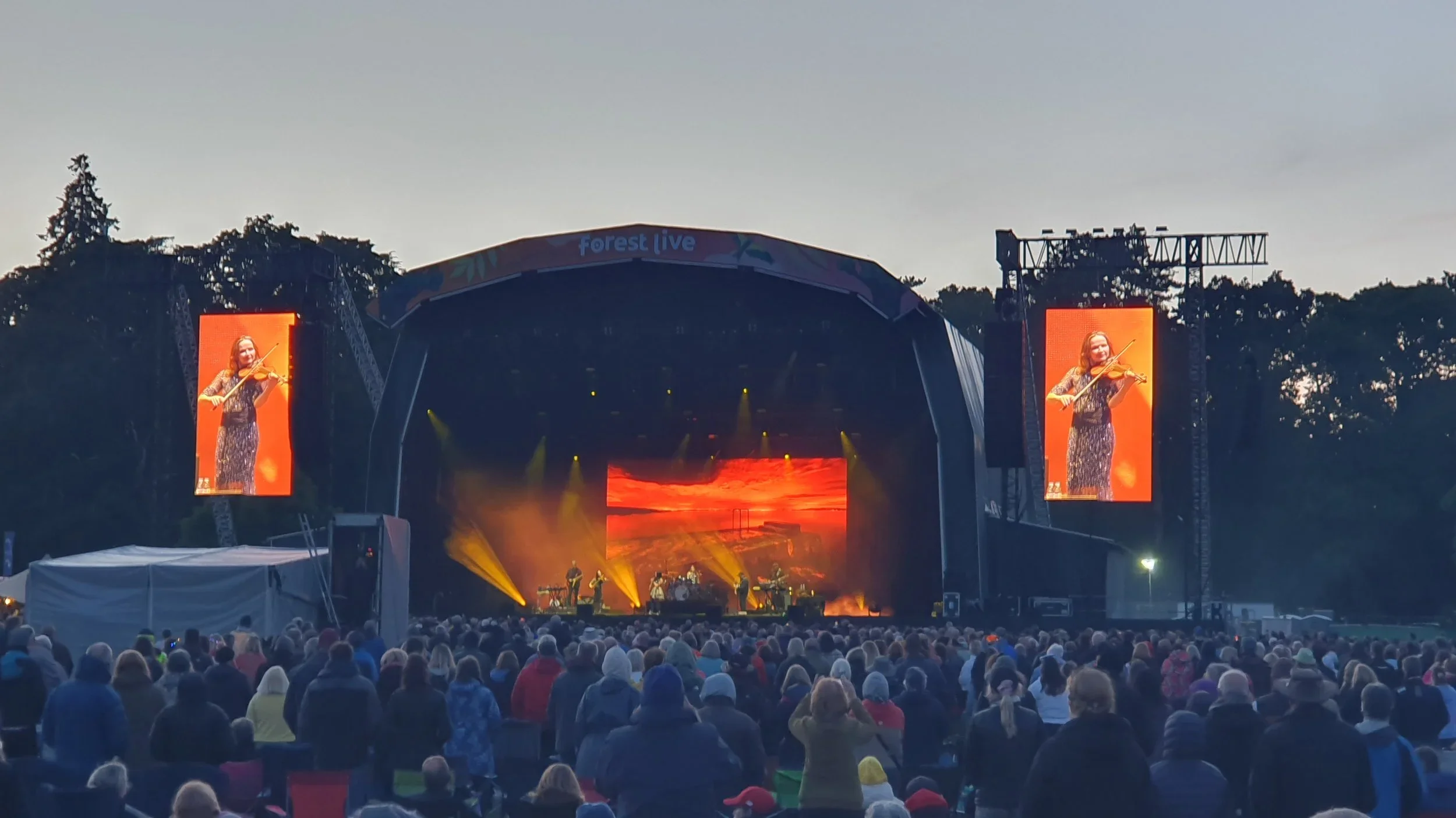Large outdoor concert stage during evening, with two large screens showing a female violinist, and a crowd of people watching.