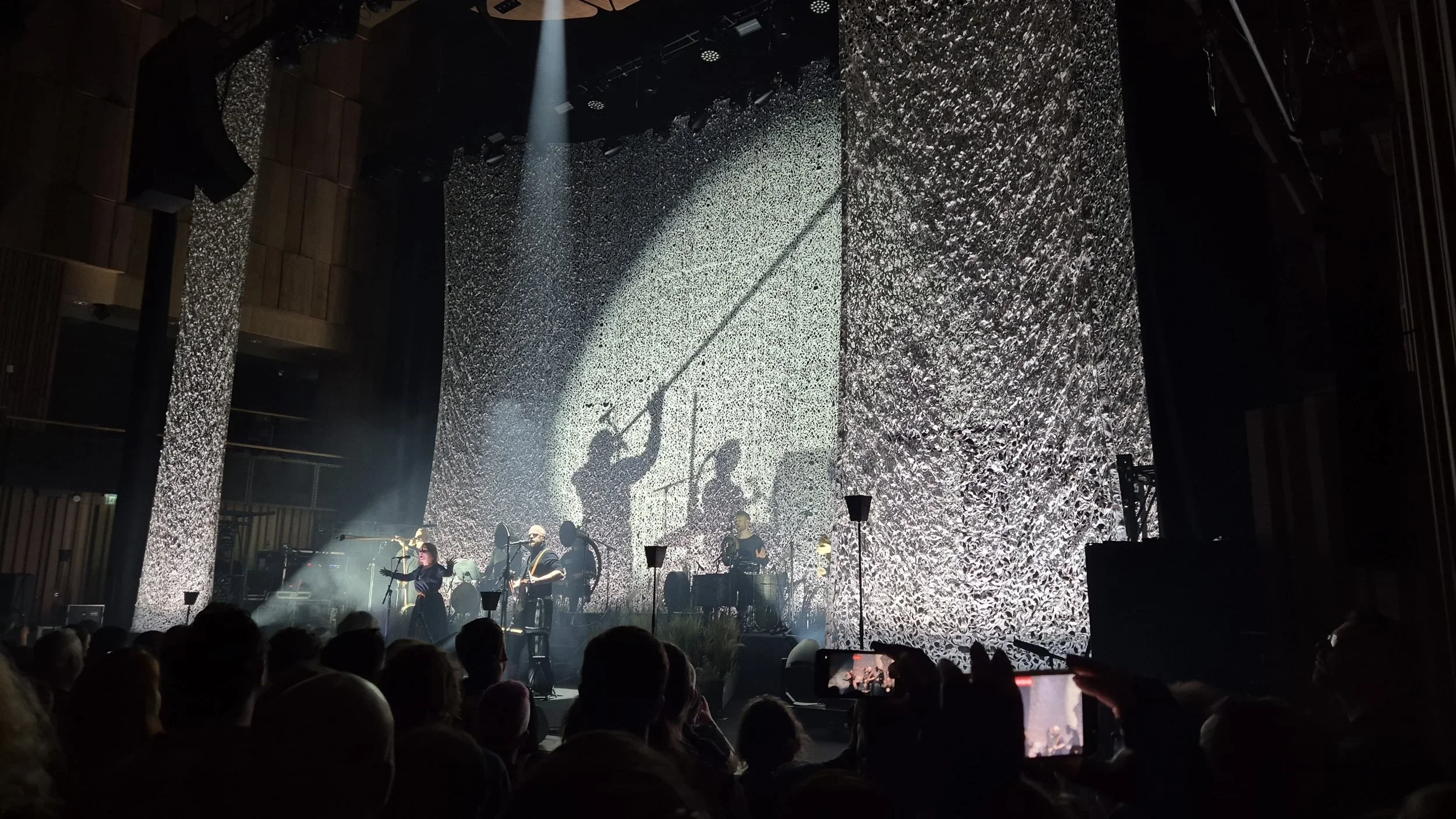A concert stage with performers and a large textured backdrop, illuminated by spotlights, and audience members in the foreground.