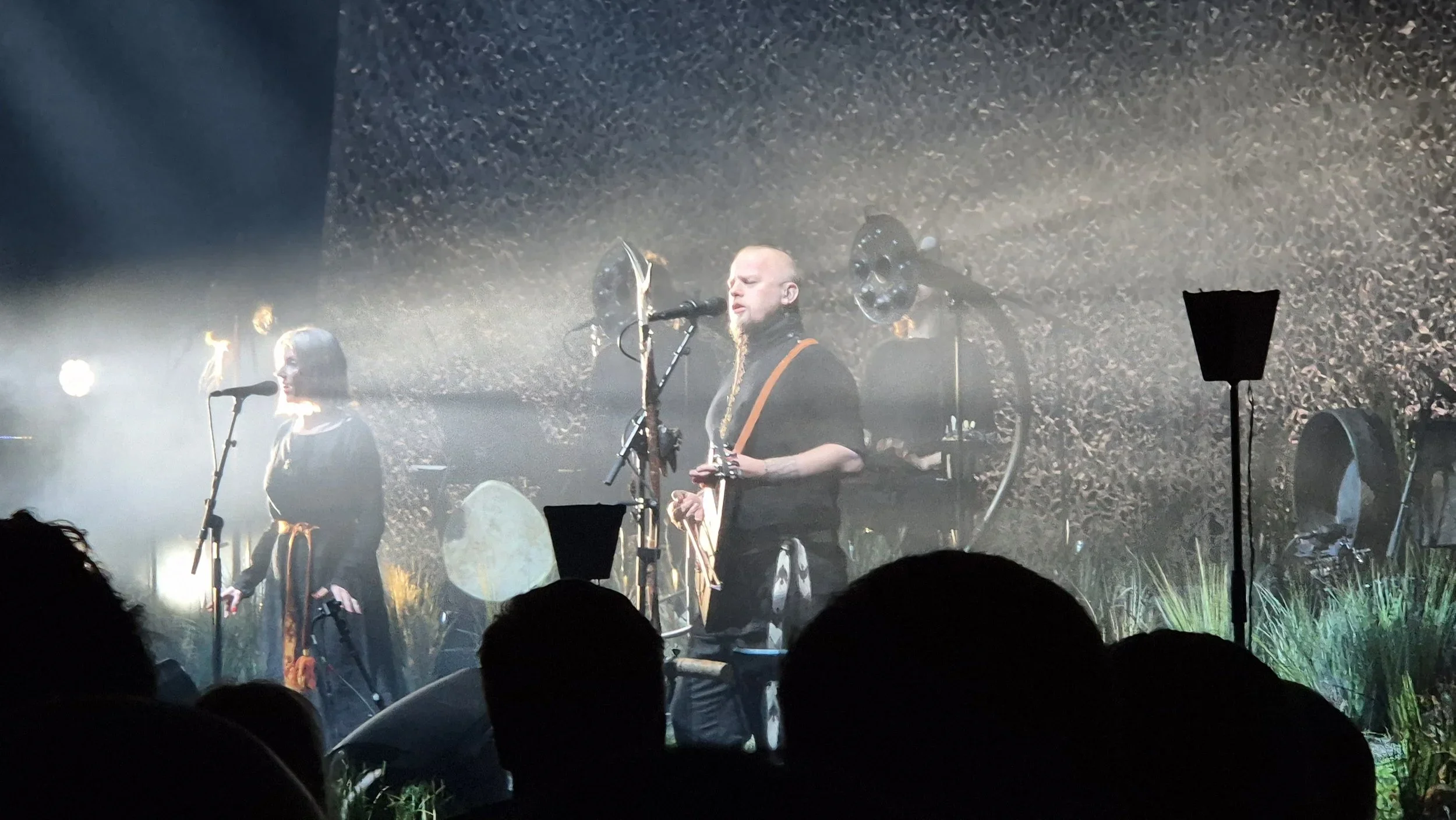 A musical performance on stage with a man singing into a microphone and playing a traditional string instrument, accompanied by a woman for backing vocals, amid atmospheric lighting and stage decorations resembling nature.