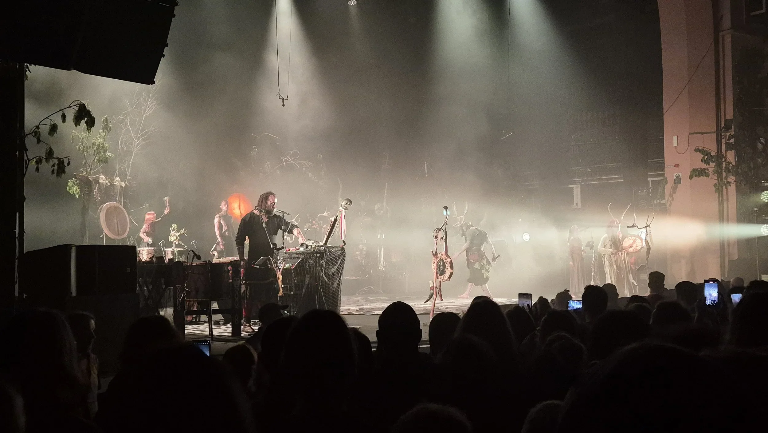 Stage performance featuring a musician with a laptop and DJ equipment on the left, performers wearing costumes and antlers on the right, and stage decor with trees and props, all illuminated by fog and stage lights.