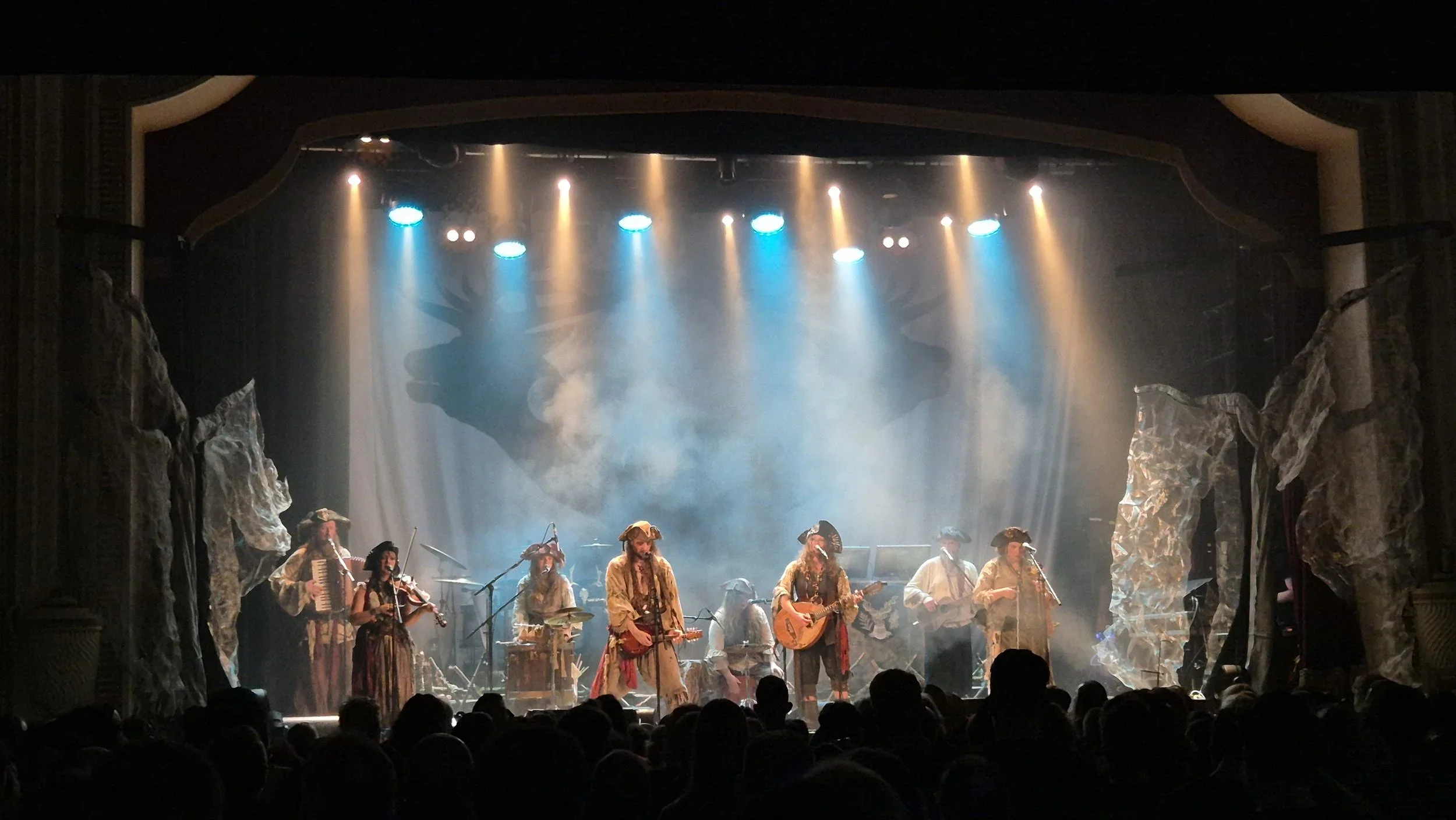 A live stage performance with musicians dressed in pirate or medieval costumes, playing instruments such as violins, guitars, and accordions, under dramatic lighting with fog effects and rocky stage decorations.