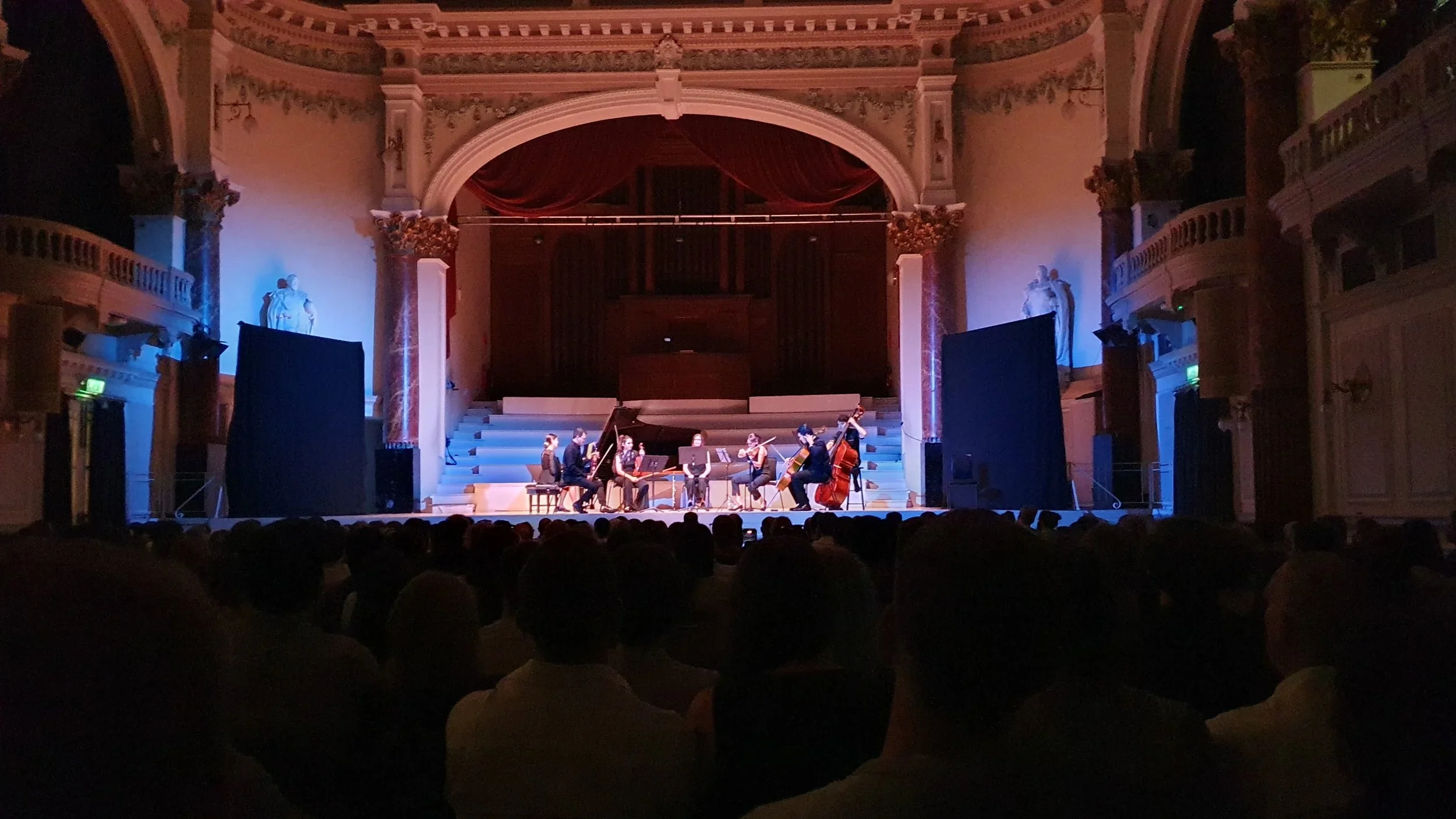 An orchestra performing on a stage in a historic concert hall, with a grand piano and a red velvet curtain, surrounded by ornate architectural details and statues.