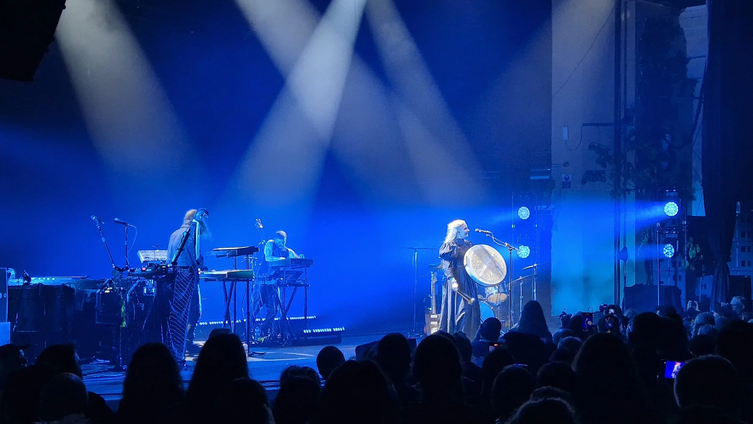 A live music performance on a stage illuminated with blue lights, featuring a female musician playing a large percussion instrument and two other musicians with electronic equipment.