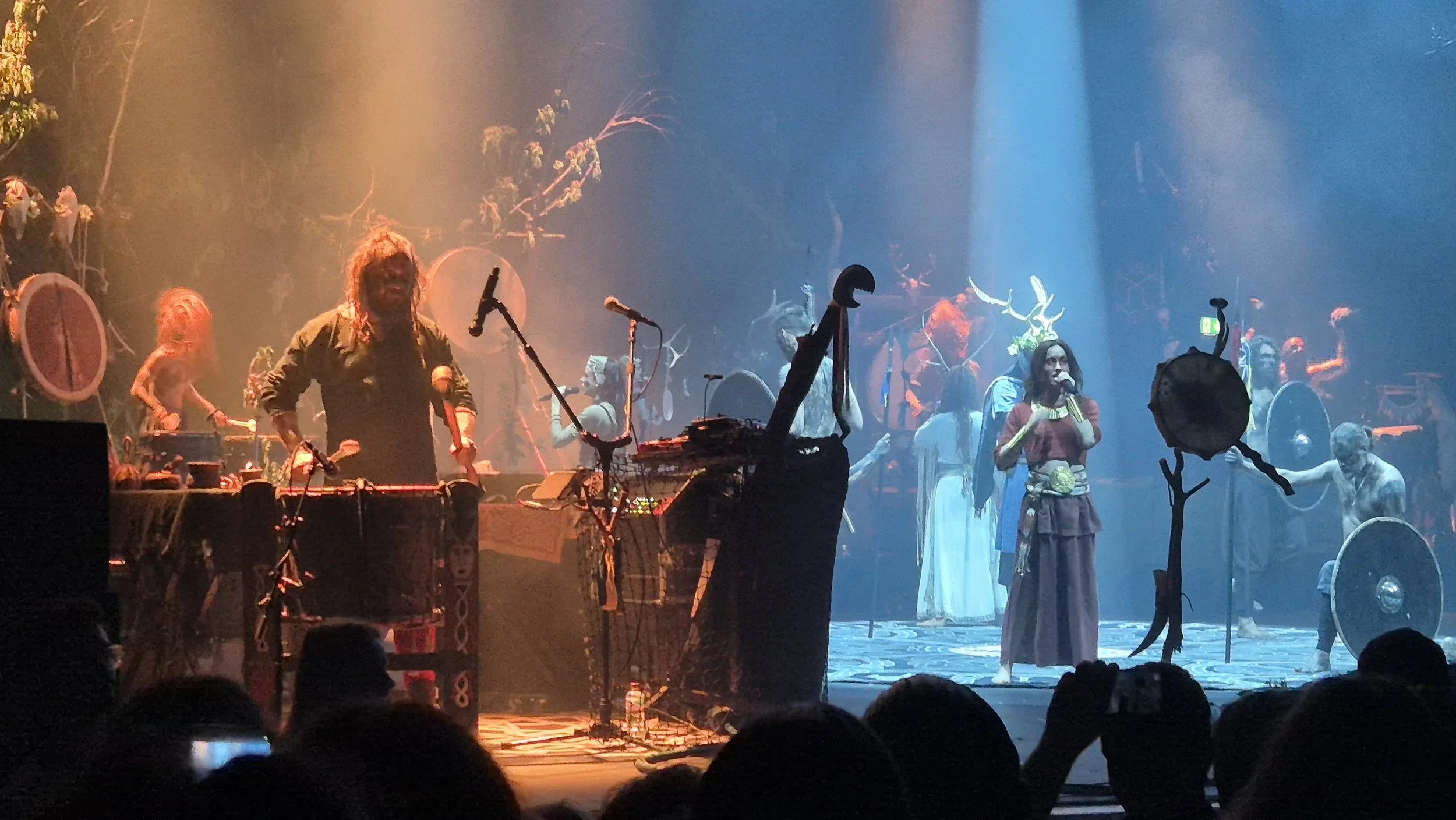 A stage performance featuring musicians and performers dressed in costumes with antler headpieces, surrounded by drums and various props, with colorful lighting creating an atmospheric scene.