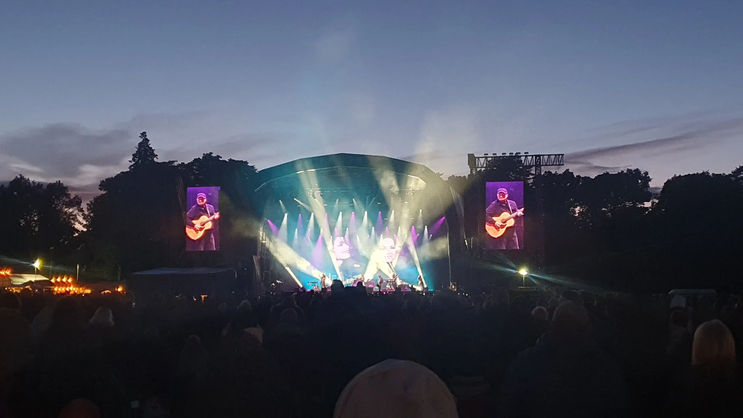 An outdoor concert stage illuminated with colorful lights during dusk, with two large screens displaying a musician playing an acoustic guitar.