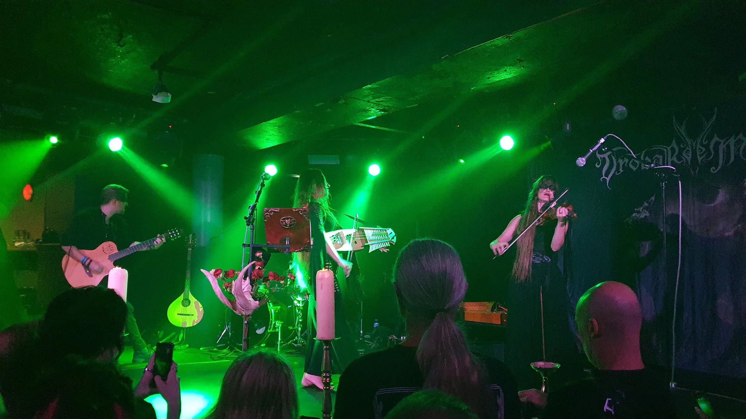 A band performing on stage with green lighting, featuring a guitarist on the left, a multi-instrumentalist in the center, and a violinist on the right, with audience members watching.