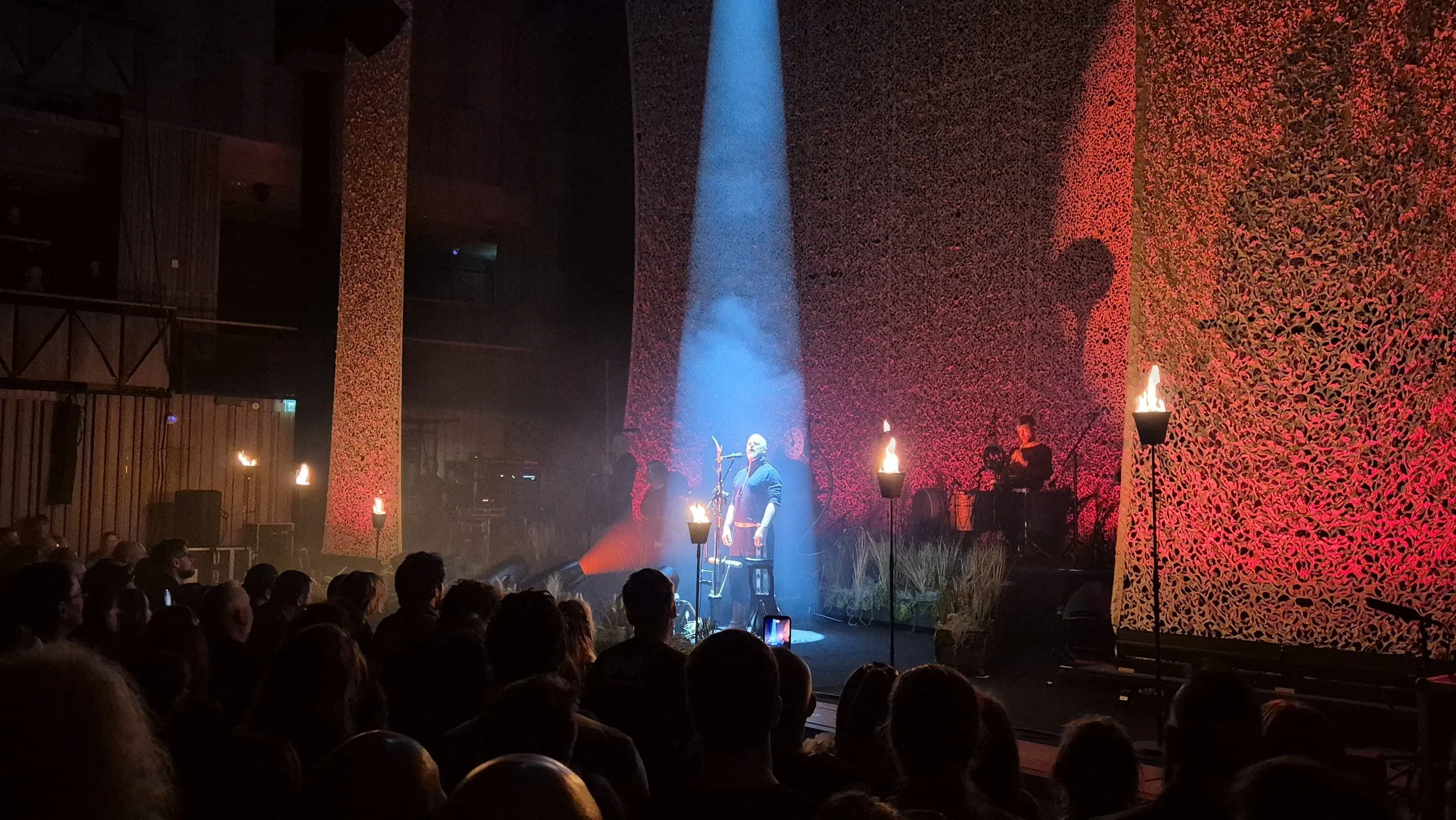 A musical performance on stage with a singer in the center, illuminated by a spotlight, and backup musicians to the right. The stage backdrop is decorated with warm-colored flames and a textured pattern, with additional lighting along the sides. The 