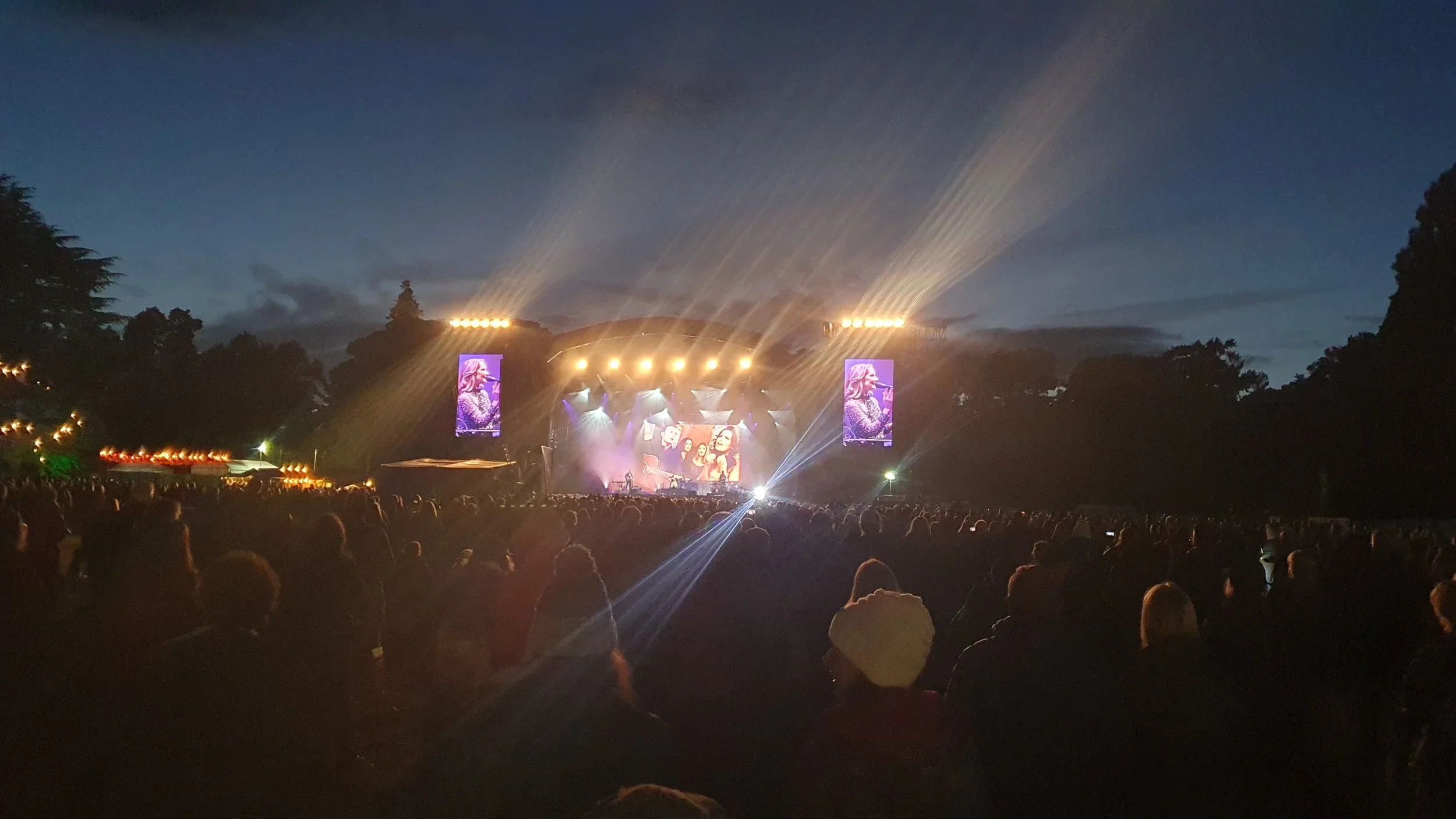 An outdoor music concert at dusk with a large crowd, stage lighting, and digital screens displaying performers, set against a darkening sky surrounded by trees.