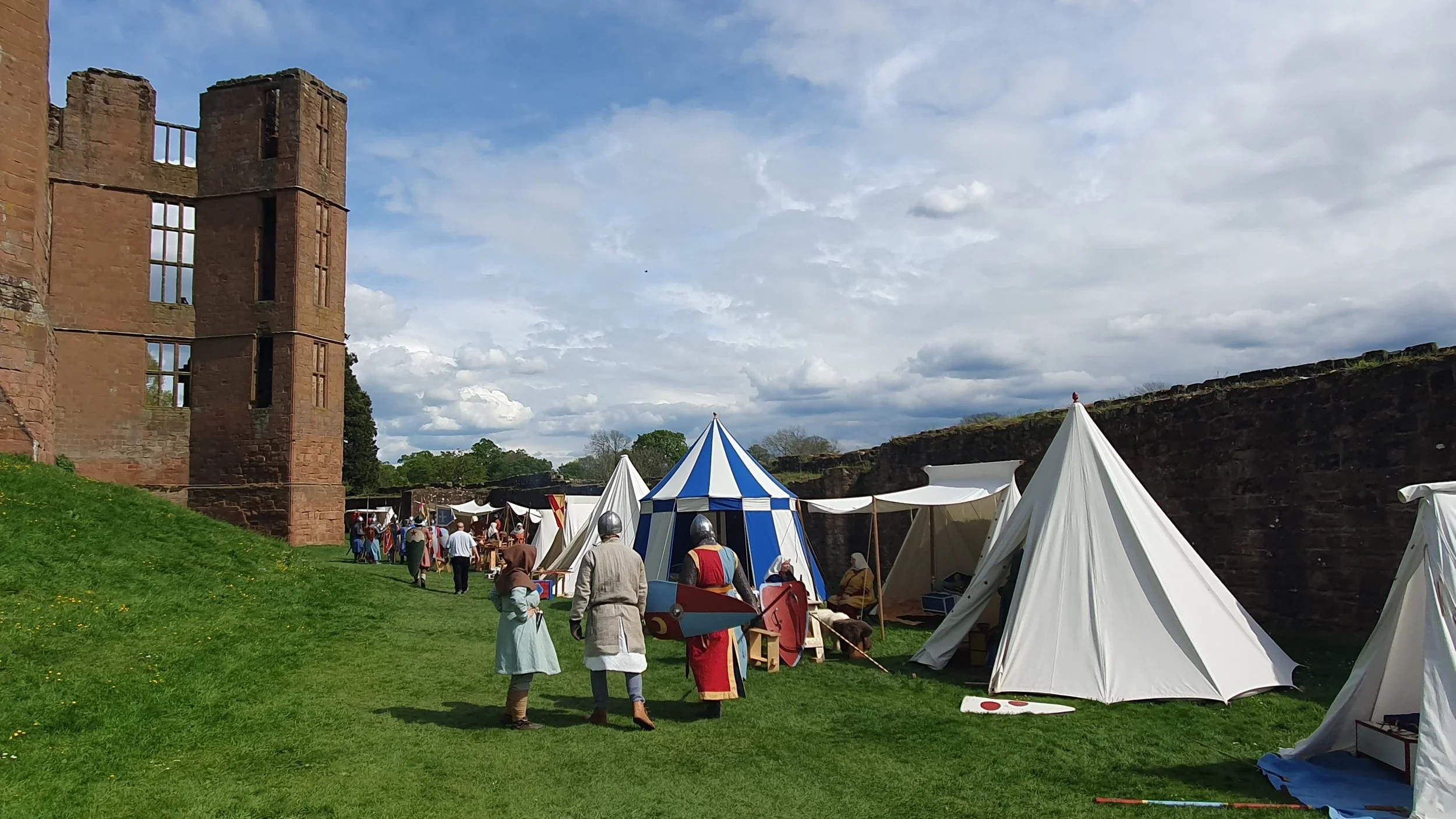 A medieval reenactment with tents, people in historical costumes, and a large brick castle wall under a partly cloudy sky.