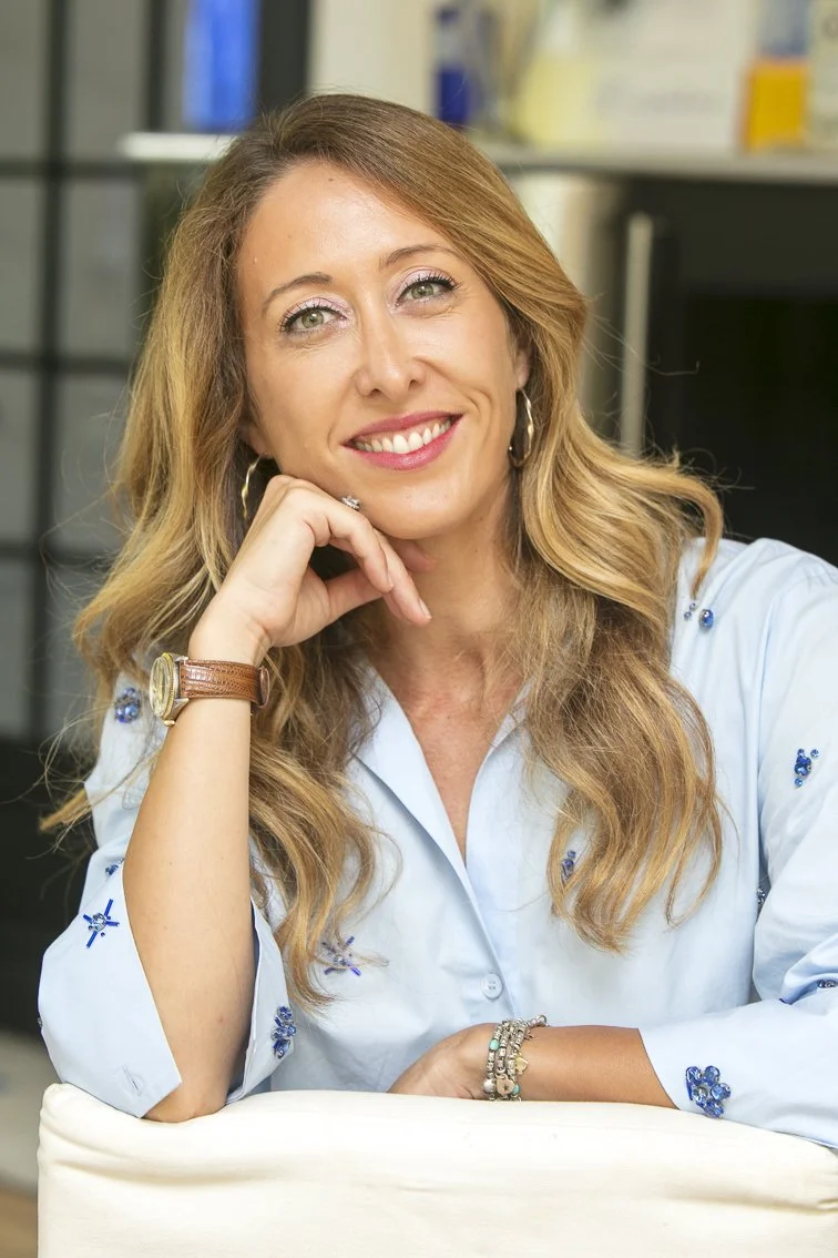 Femme souriante aux cheveux longs, portant des boucles d'oreilles et une montre en bracelet, assise à une table dans un environnement intérieur, probablement un bureau.