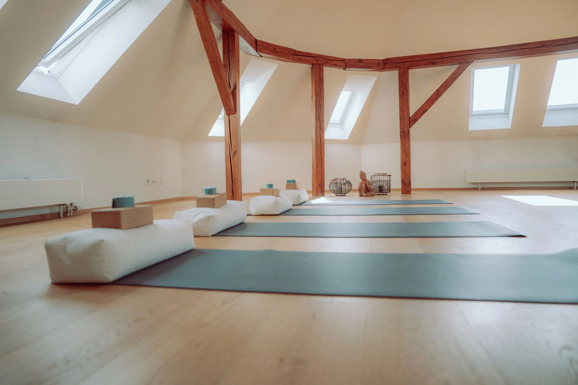 Yoga-Raum mit Matten, Meditationskissen und Dekorationen, beleuchtet durch Dachfenster mit Holzträgern.
