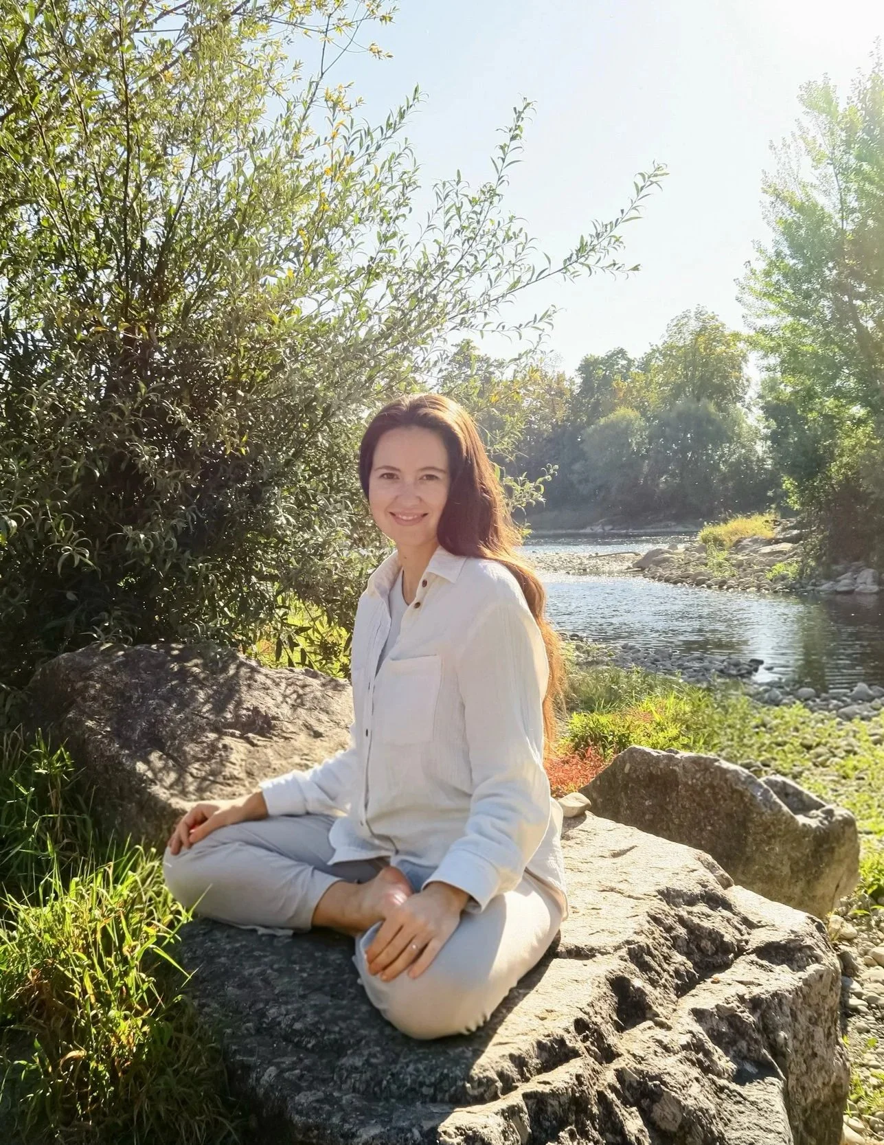 Eine Frau sitzt entspannt auf einem großen Stein am Flussufer, umgeben von Bäumen und Natur, bei sonnigem Wetter.