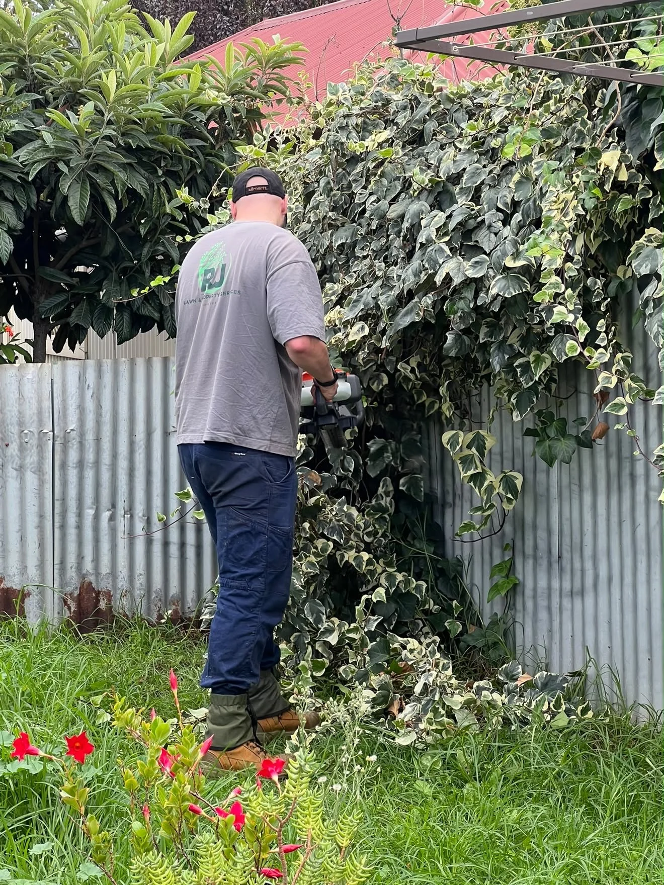 Person using a grass trimmer in a backyard with tools and plants, surrounded by trees and a steel fence.
