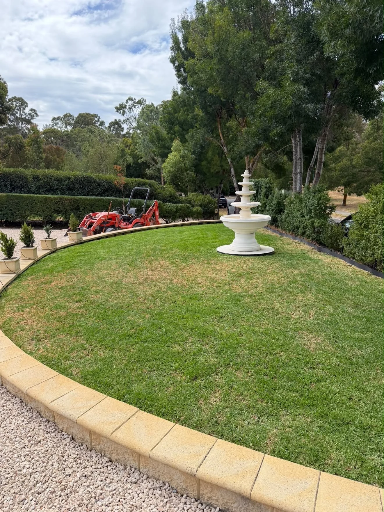 A well-maintained garden lawn with a white decorative fountain, potted plants lining the edge, and surrounded by trees and hedges on a partly cloudy day.