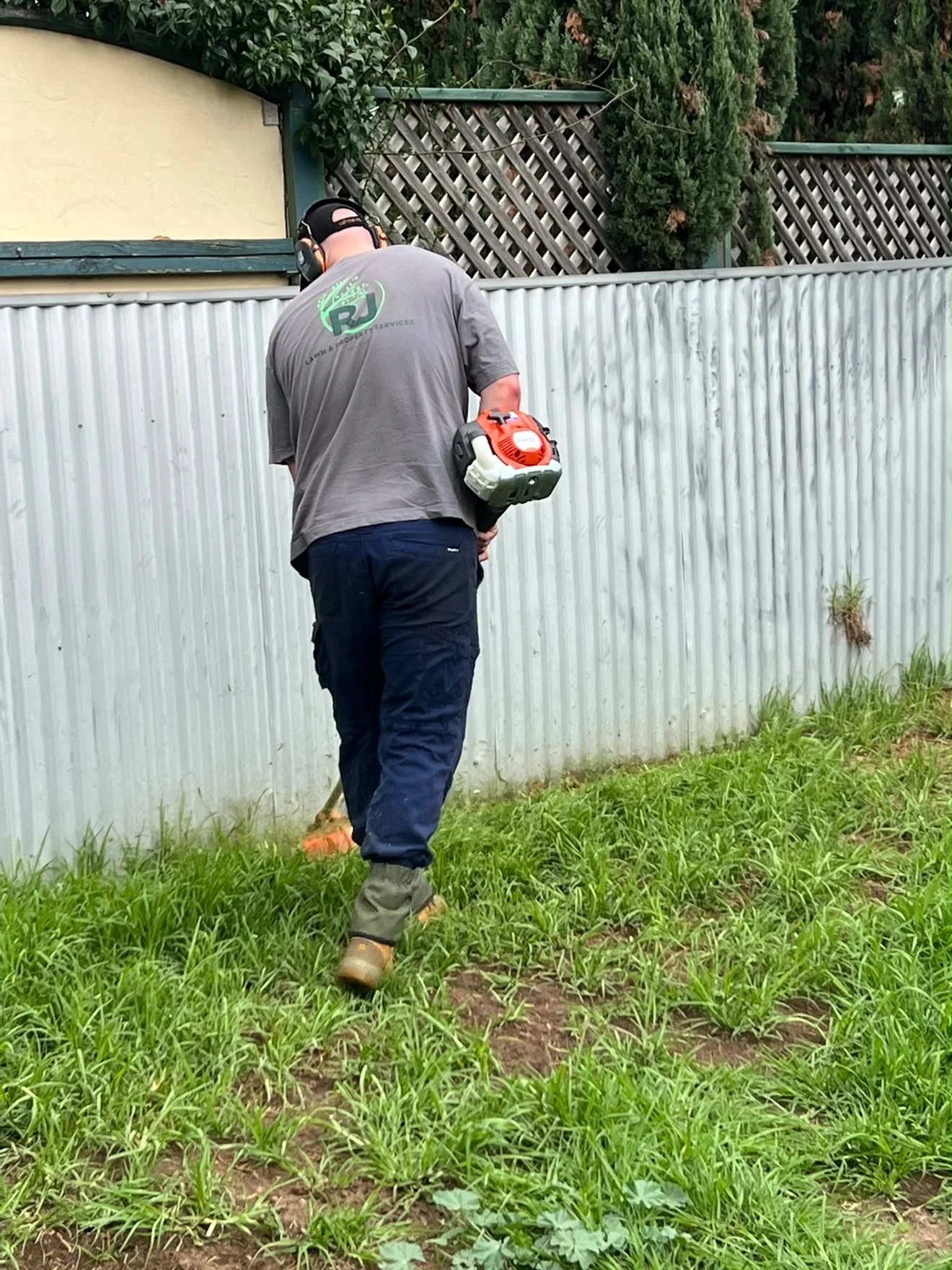 A person wearing a gray t-shirt, dark pants, and boots is using a weed trimmer along a metal fence in a backyard. The person is wearing a black cap and headphones. There is greenery and trees in the background.