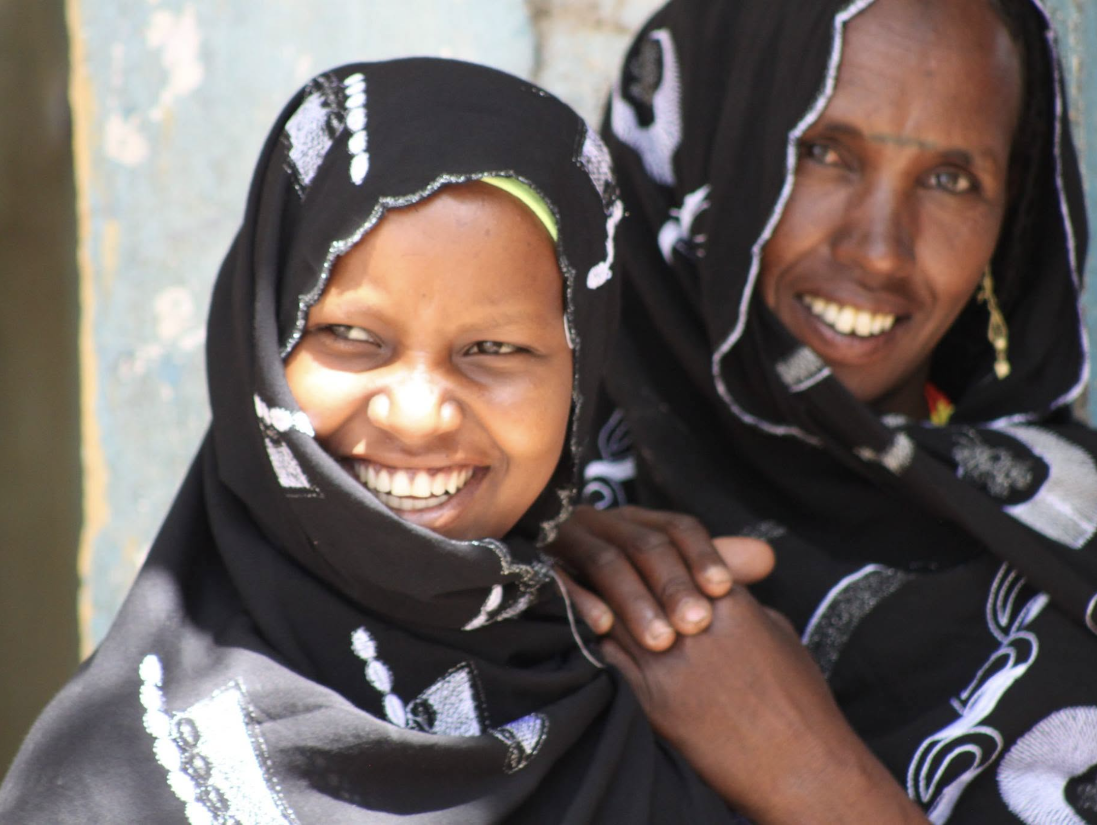 Two smiling women wearing black headscarves with white embroidery, one woman is sitting with hands on her lap, the other is leaning close to her.