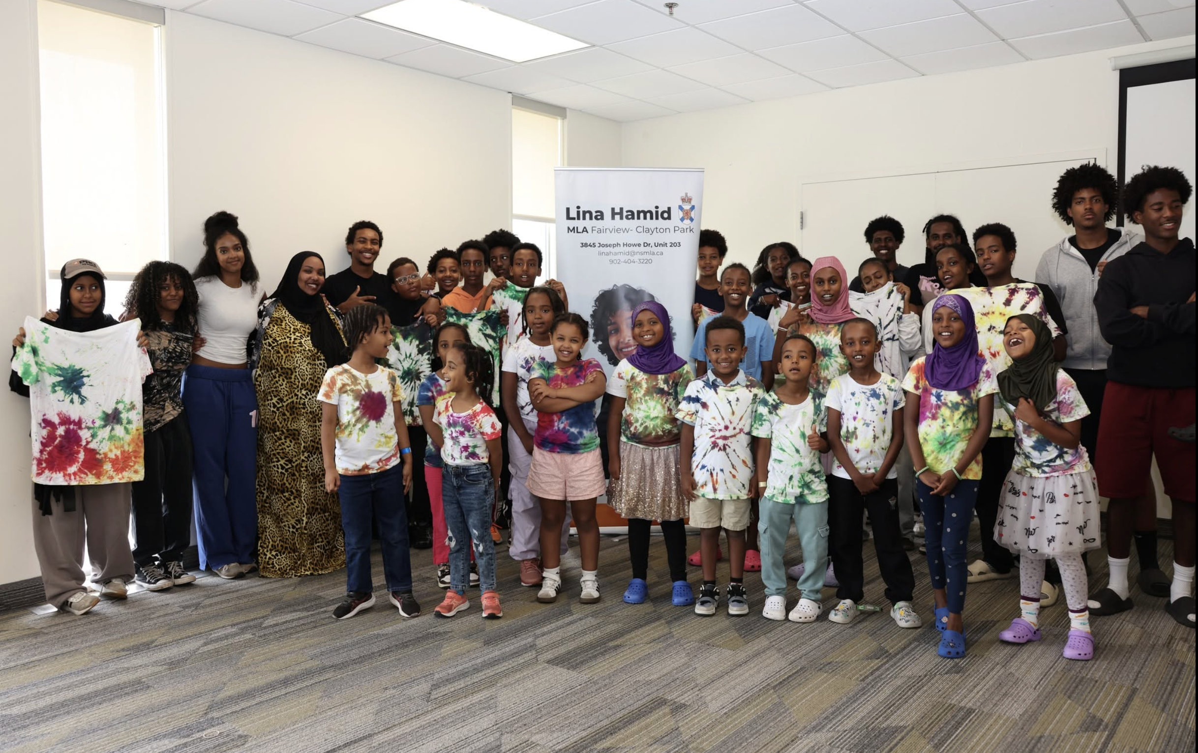 A group of children and teenagers, some smiling and wearing tie-dye shirts, standing in a room with light walls and a banner for Lina Hamid, MLA Fairview-Clayton Park, with contact details.