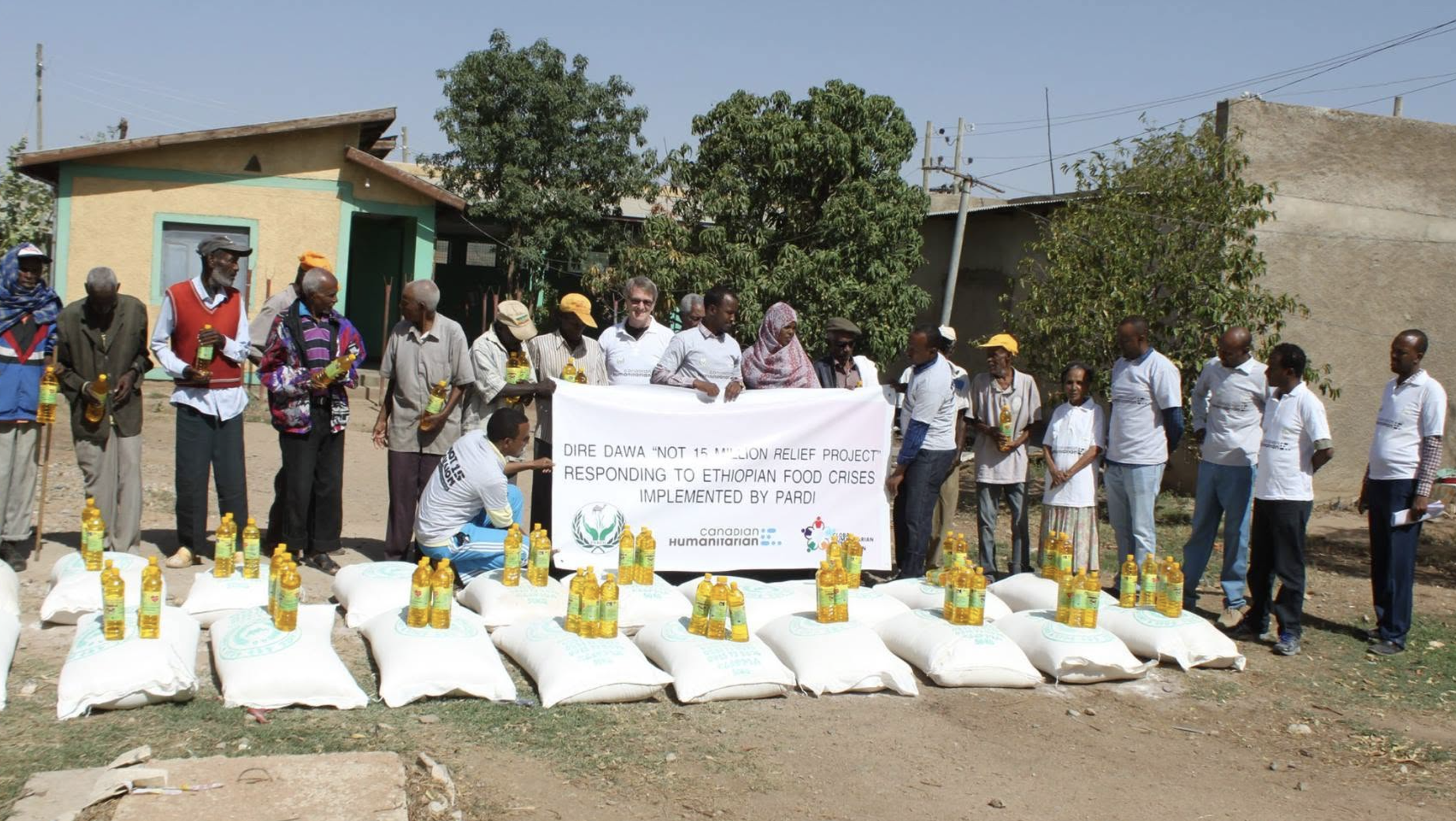 A group of people standing outdoors in front of a banner and supplies, participating in a relief project to respond to Ethiopian food crises, with bottles of cooking oil and large bags of flour or grain on the ground.