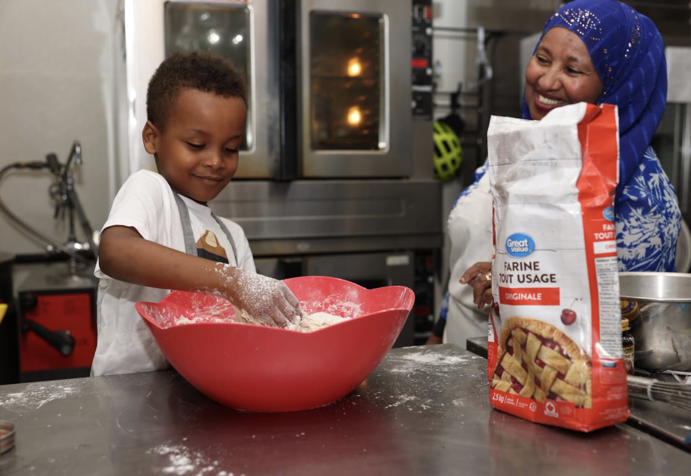 A young boy and an older woman baking together in a kitchen. The boy is mixing dough in a red bowl, and the woman is smiling while holding a bag of flour. There are kitchen appliances and ingredients visible in the background.