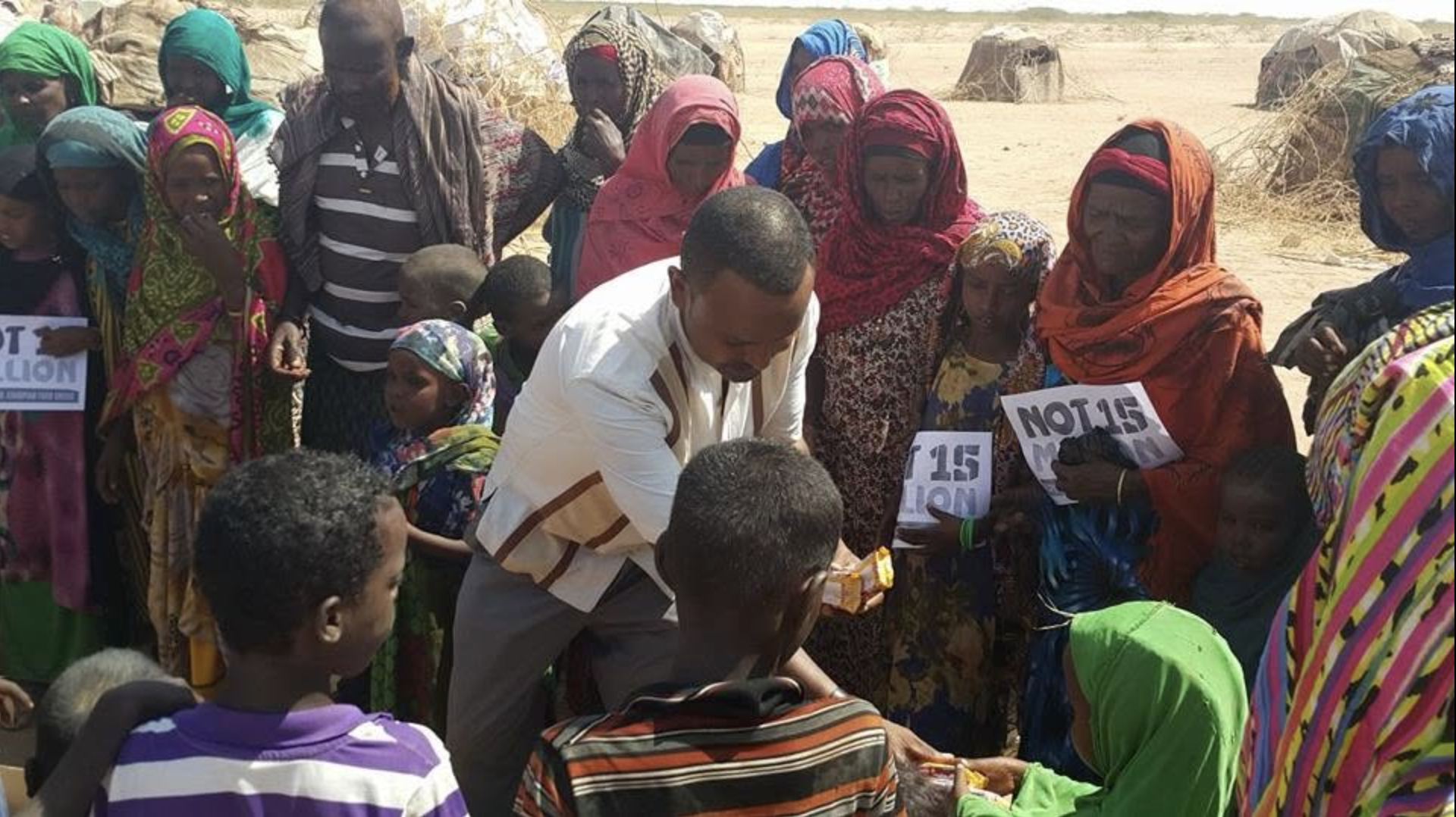 A group of people gathered outdoors in a desert-like area, with many women wearing colorful headscarves and holding signs that say 'NOT 15 MILLION'. A man in a white shirt is handing something to a seated person.