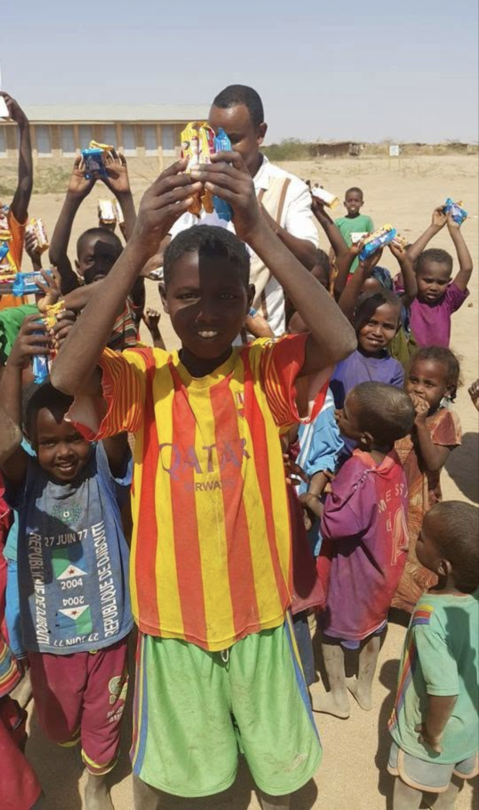 A group of children in a sandy, open area holding small gift packages, with one boy in the foreground wearing a red and yellow striped sports jersey, lifting a gift in the air.