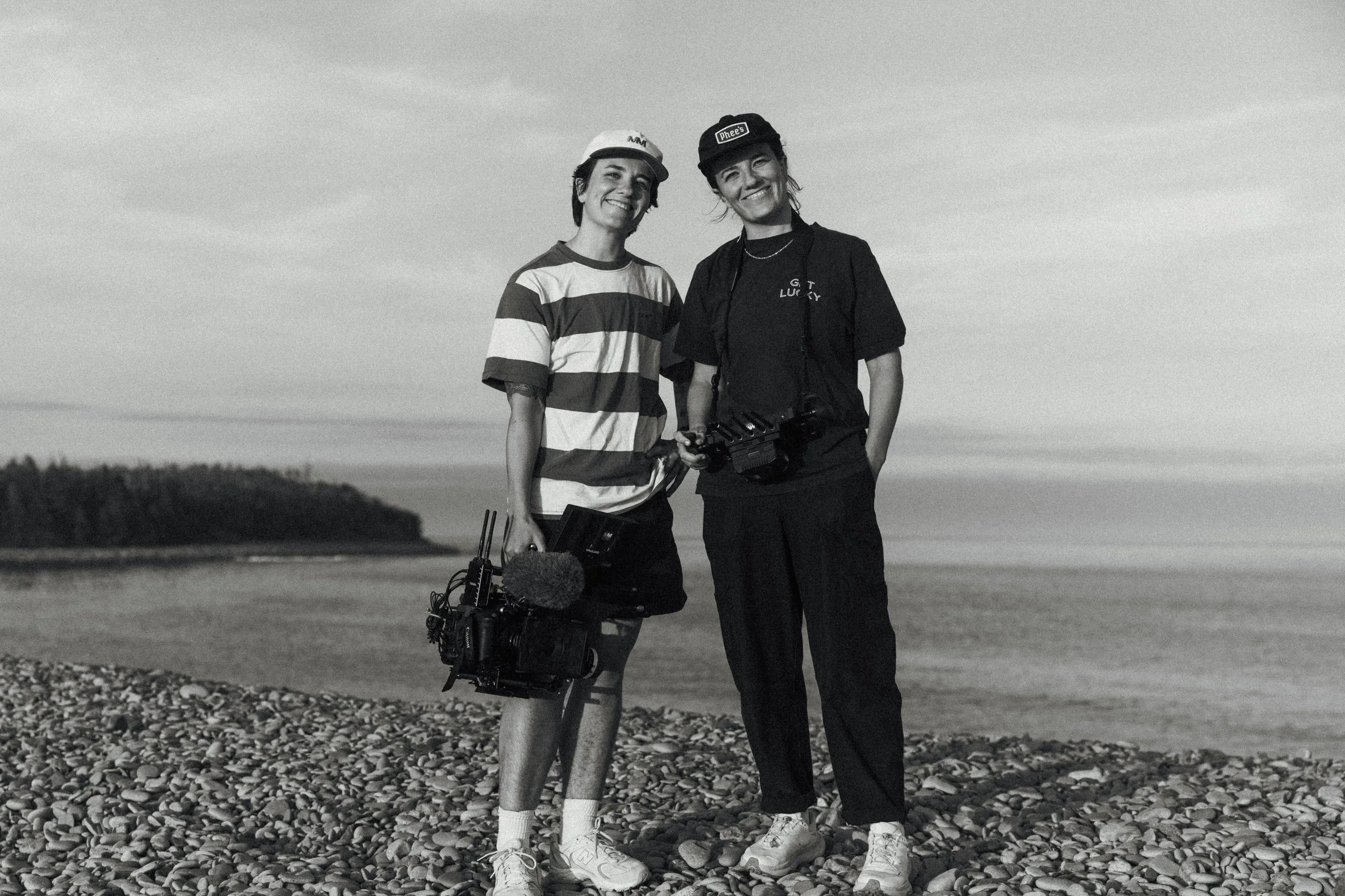 Queer twin siblings standing on a rocky beach, smiling, with a body of water and cloudy sky in the background. One woman is holding a camera and a microphone and they own a video production company