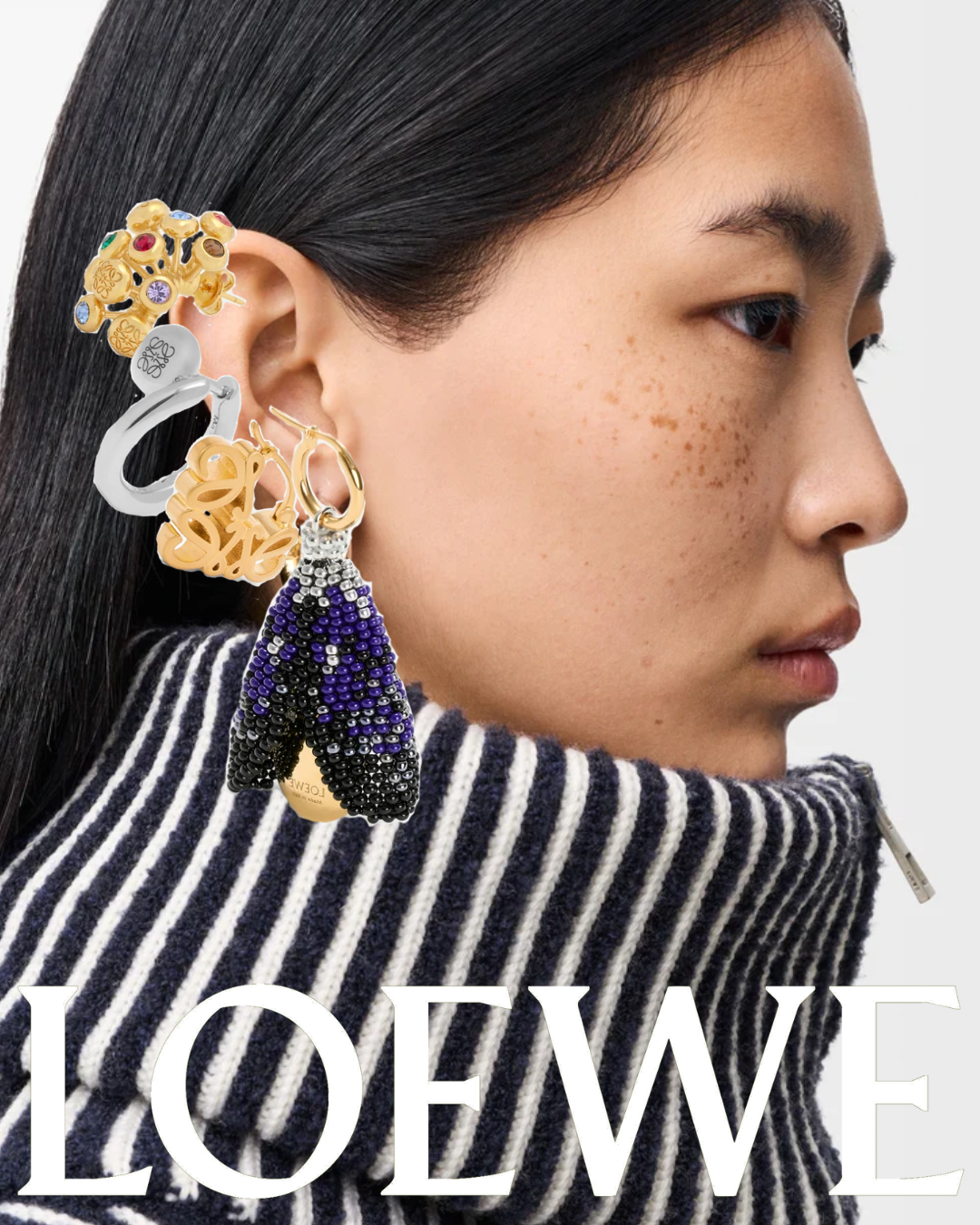 Close-up profile of a woman with dark hair and freckles, wearing a striped black and white turtleneck, adorned with multiple earrings including gold, silver, and beaded designs, against a plain background with the word 'LOEWE' at the bottom.