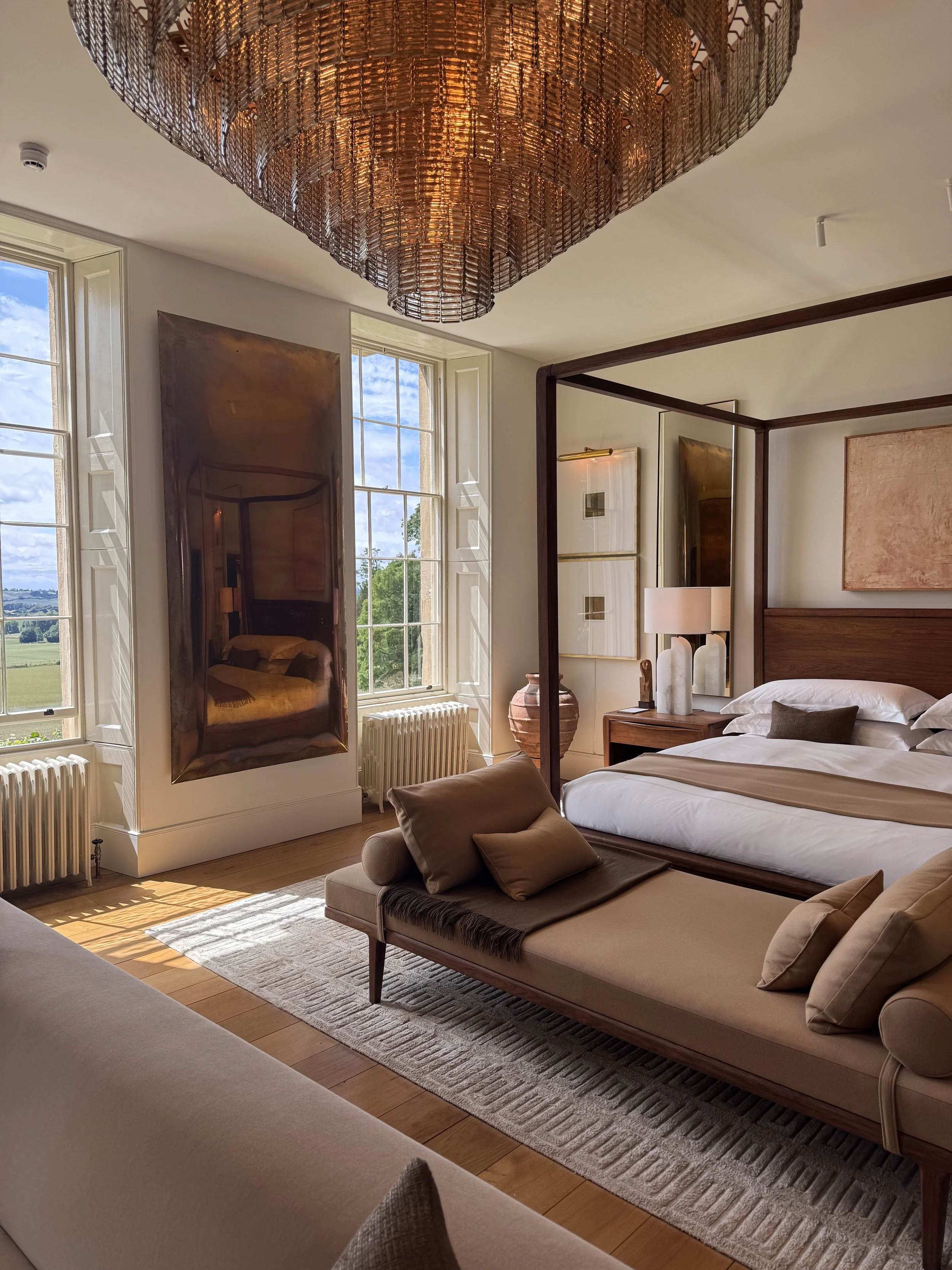 Interior of a bright bedroom with large windows, a bed with a wooden headboard, a beige chaise lounge with pillows, an abstract painting, a large decorative mirror, and a chandelier hanging from the ceiling.