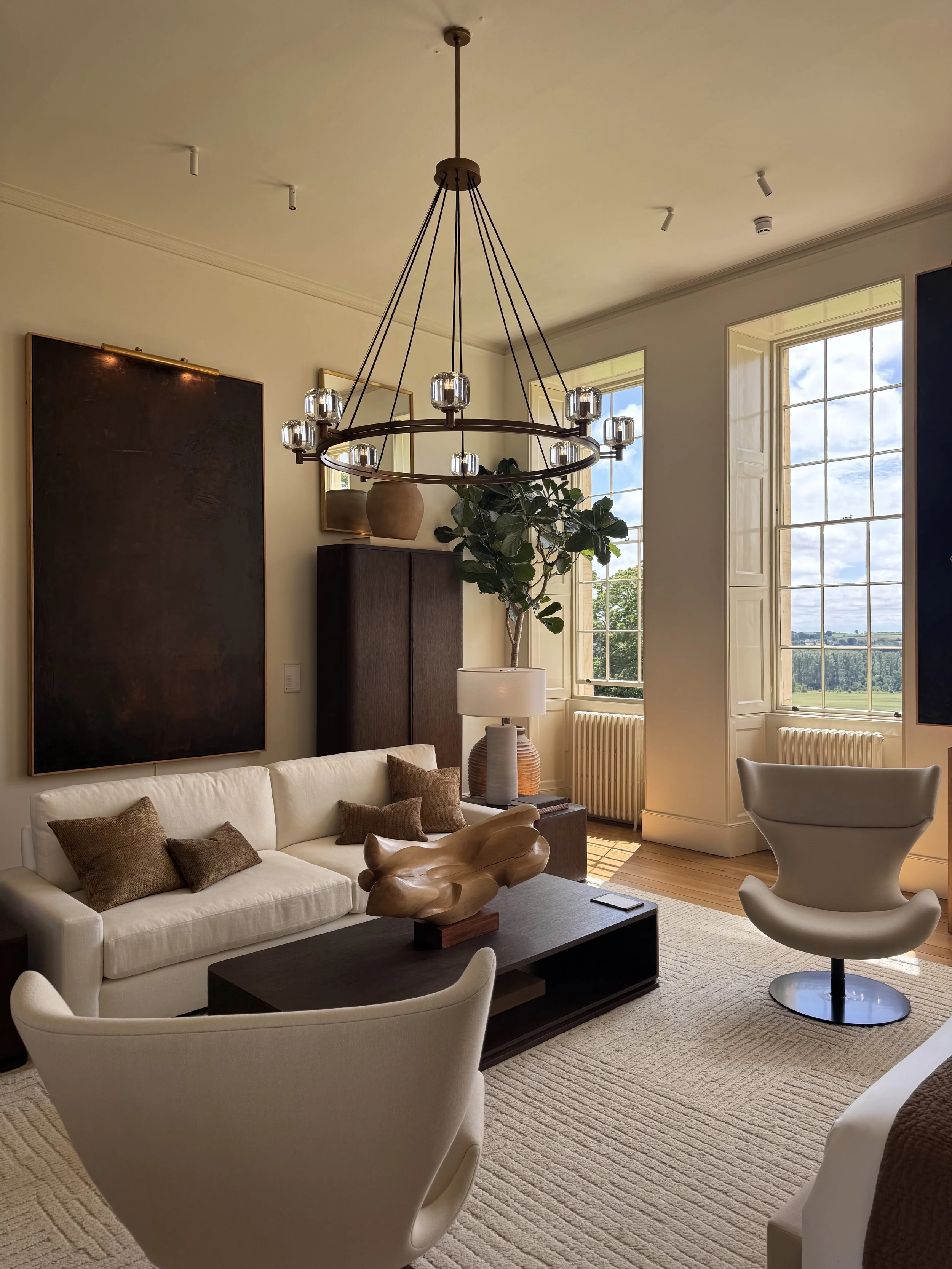 Living room with a cream sofa, two cream armchairs, a dark coffee table with a wooden sculpture, wall art, a large black cabinet, potted plant, table lamp, large windows showing a scenic outdoor view, and a modern chandelier.