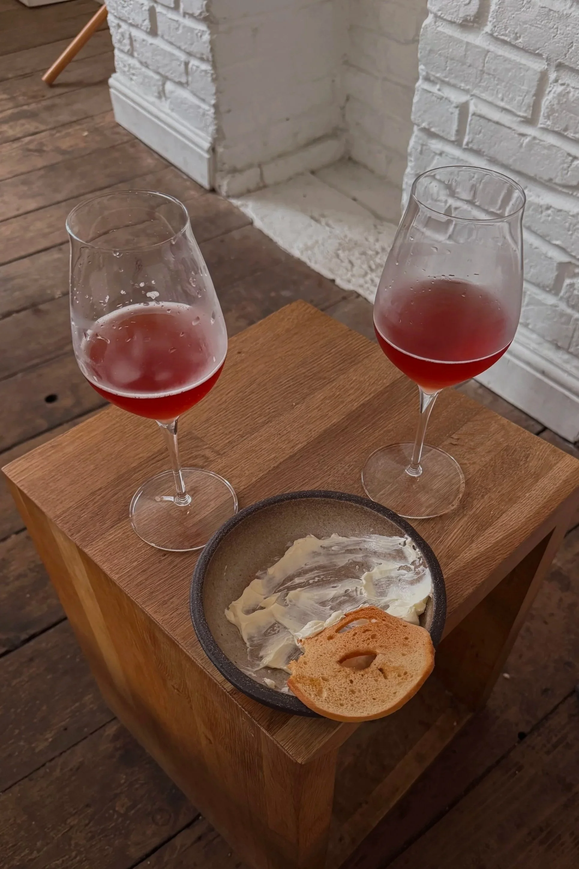 Two glasses of pink wine on a wooden table near a bowl of spread with bread and toast, in front of a white brick fireplace on a wooden floor.