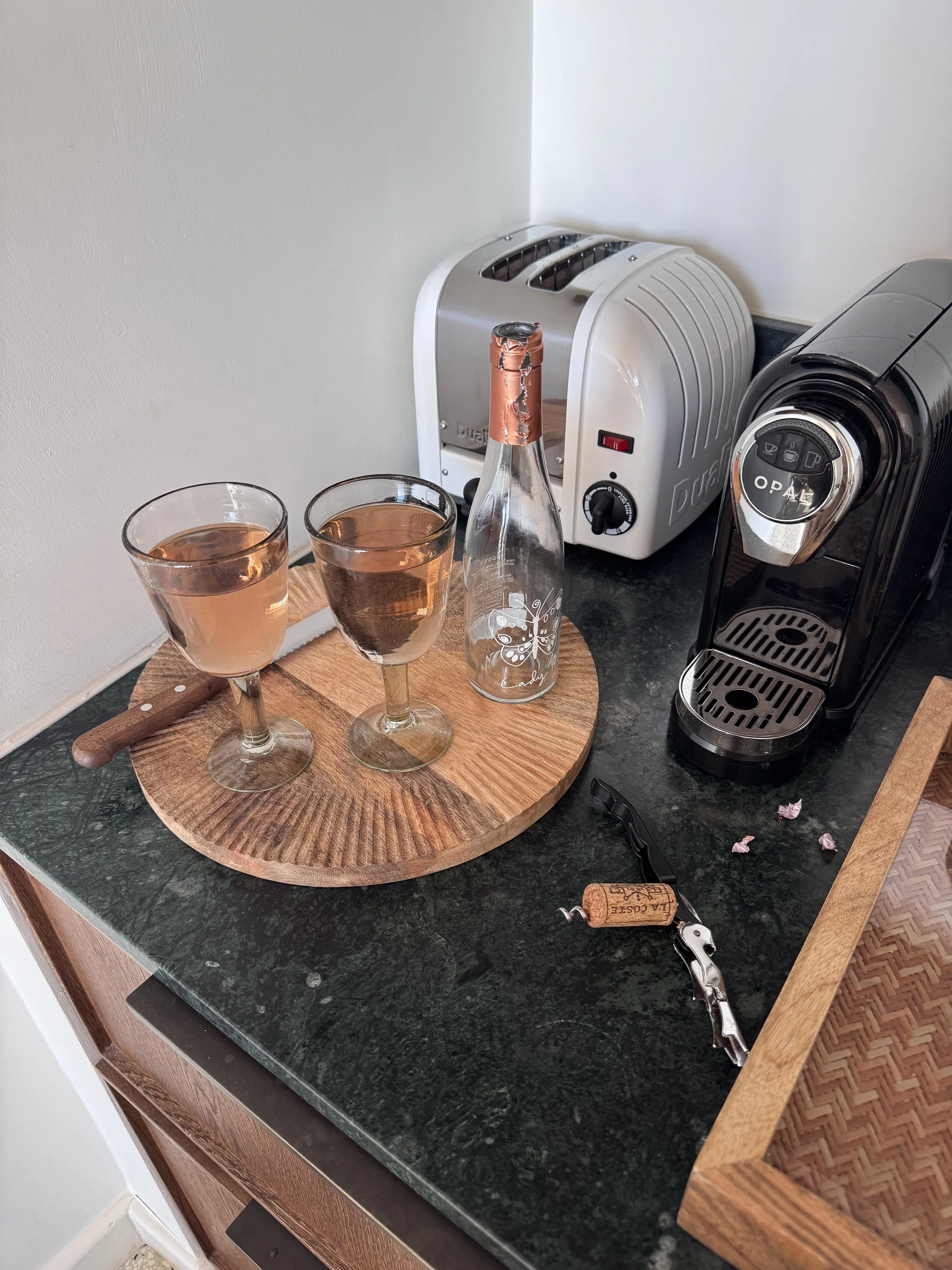 Two glasses of rosé wine, a bottle with a butterfly design, a corkscrew, a white toaster, and a black espresso machine on a dark green marble countertop in a kitchen.