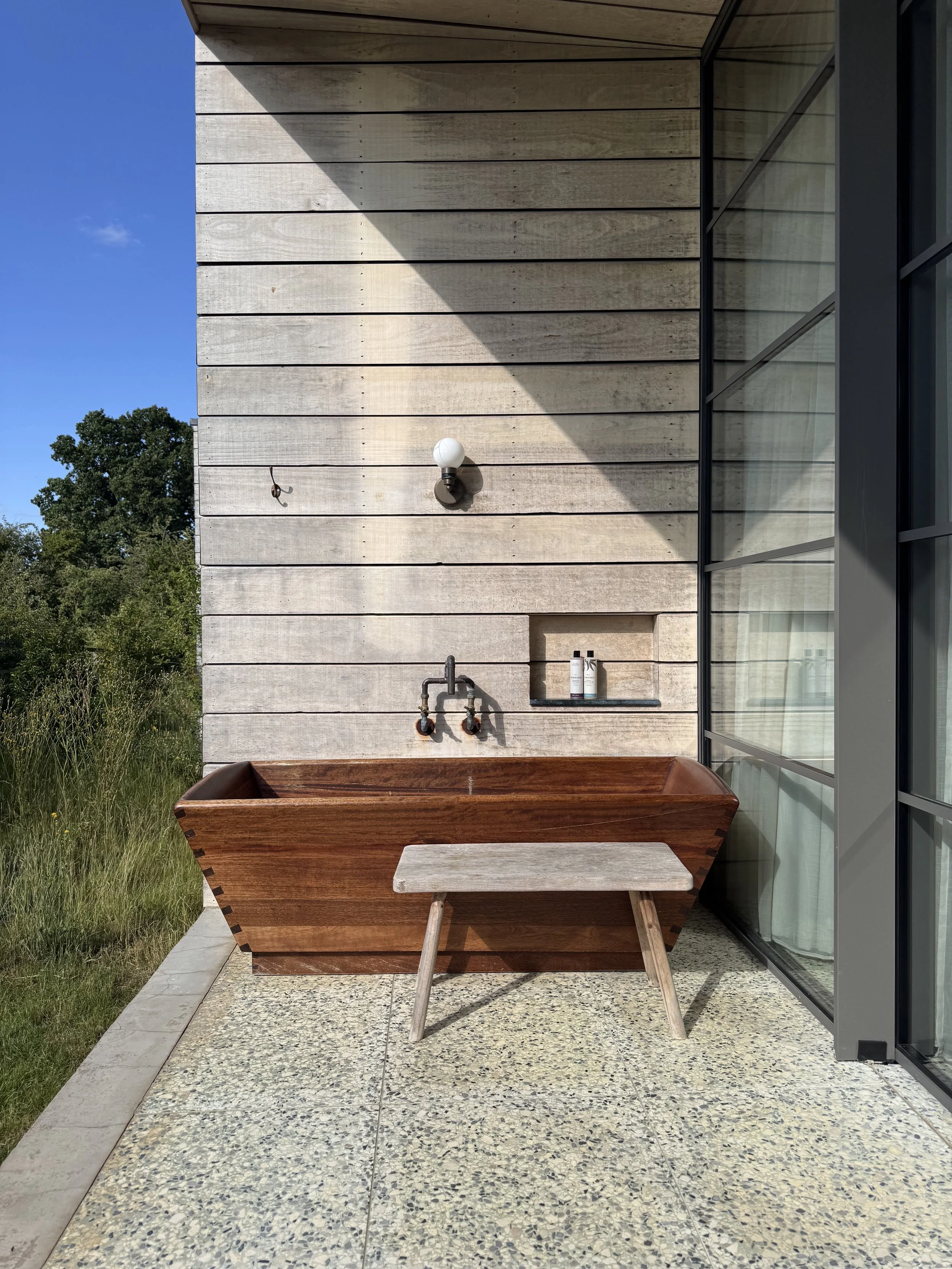 Outdoor patio with a wooden bathtub, small bench, a wall-mounted white globe light, and two bottles of skincare products, with a glass building and greenery in the background.