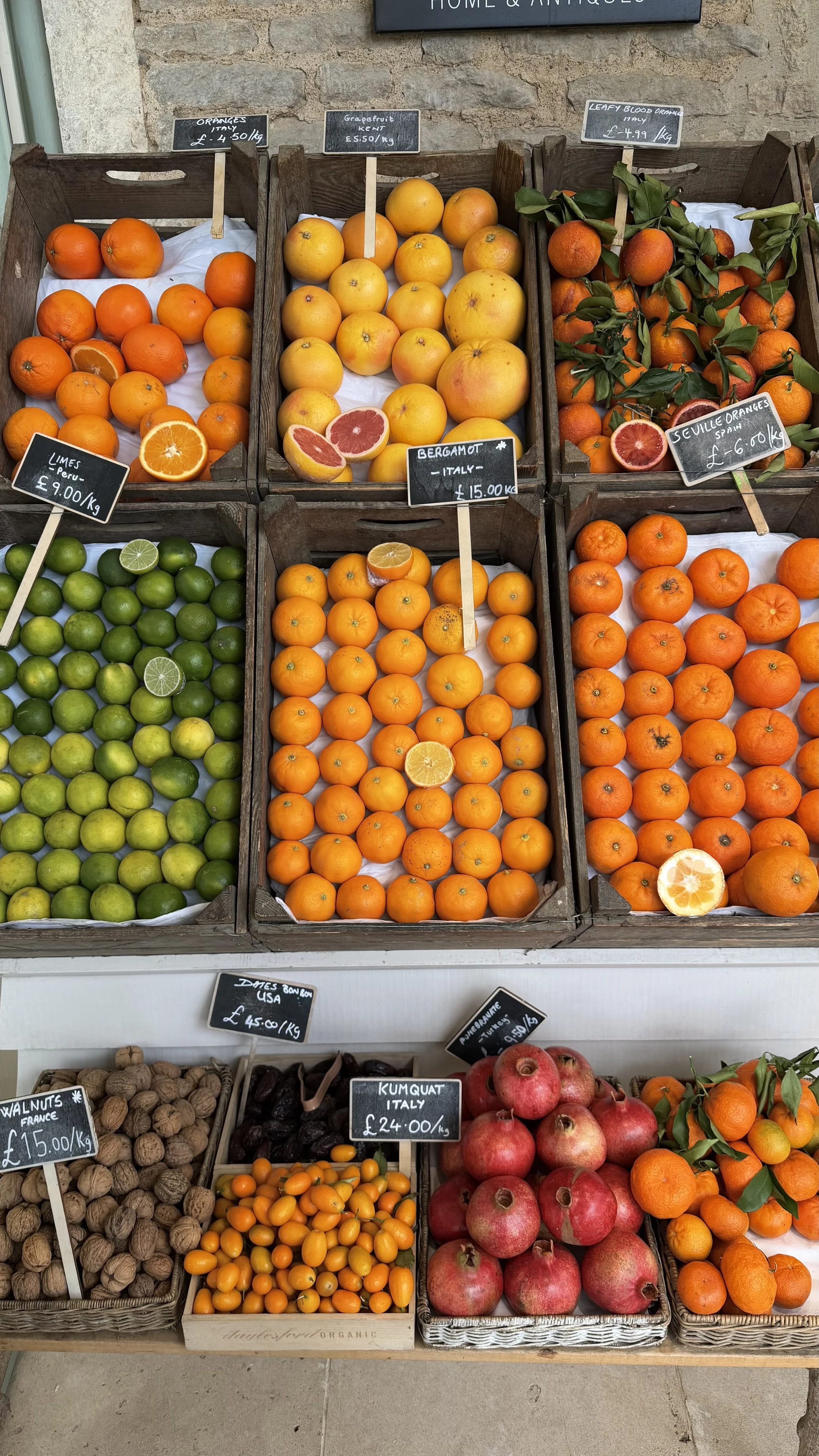 Display of various citrus fruits and other produce in wooden baskets at a market. From left to right, top to bottom: oranges, grapefruits, blood oranges with leaves, limes, tangerines, pomegranates, walnuts, dates, kumquat, pomegranates, and oranges.