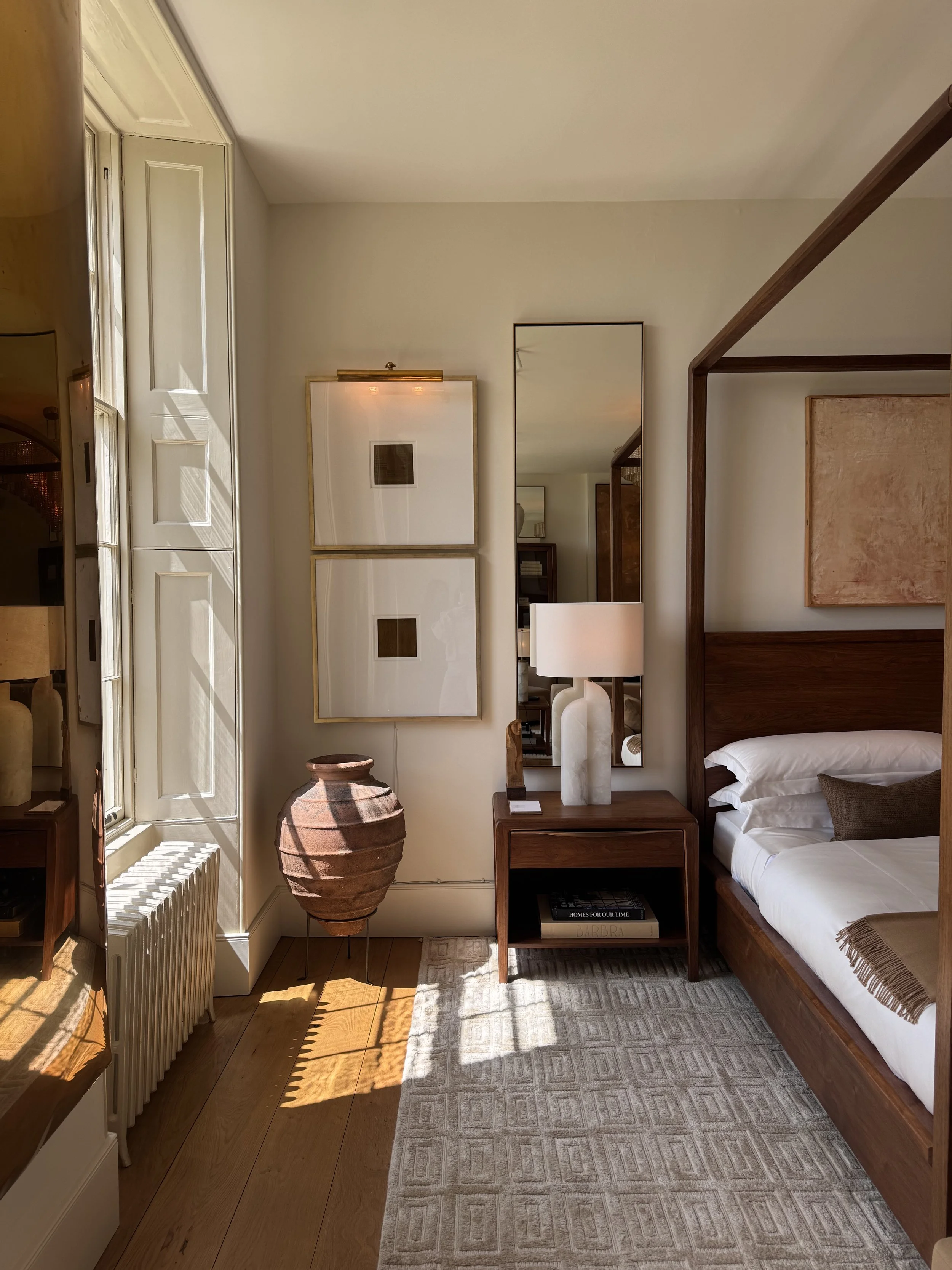 A cozy bedroom corner with a bed, nightstand, mirror, and artwork. Natural light enters through a window, illuminating wooden flooring and a patterned rug.